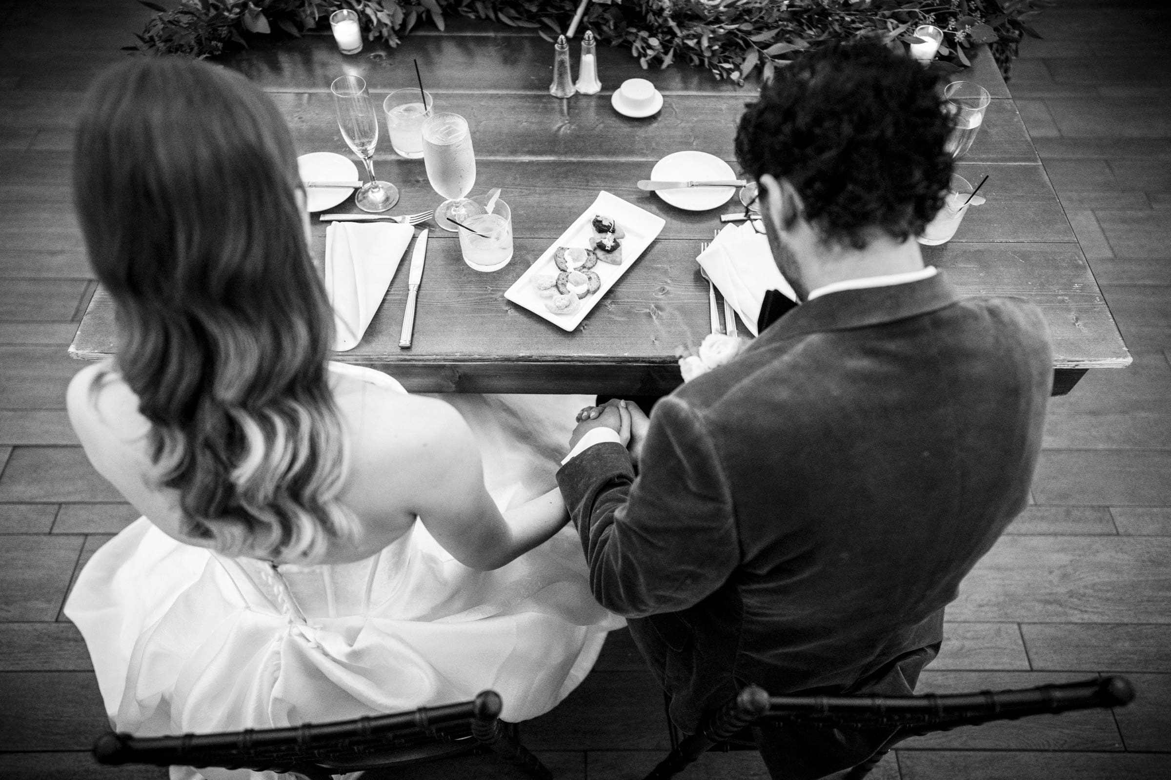 A bride and groom sit side by side at a wooden table, holding hands, viewed from above. The table is set for a meal, and elegant desserts are placed in the center. The scene is black and white.