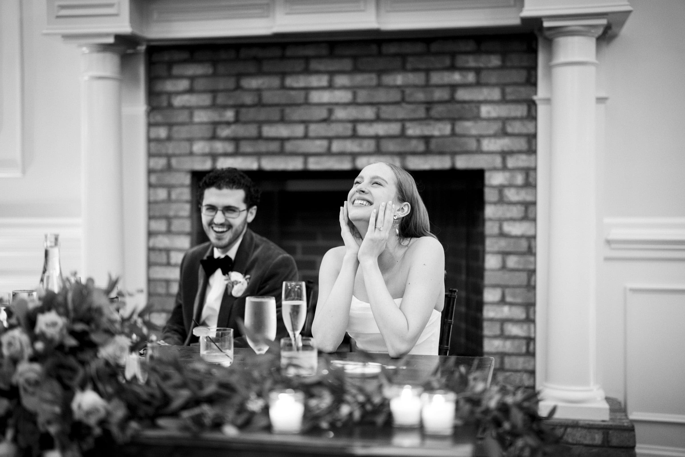 A smiling couple sits at a table in front of a brick fireplace; the woman rests her chin on her hands, looking joyful, while the man next to her also smiles. The table is decorated with flowers and candles.