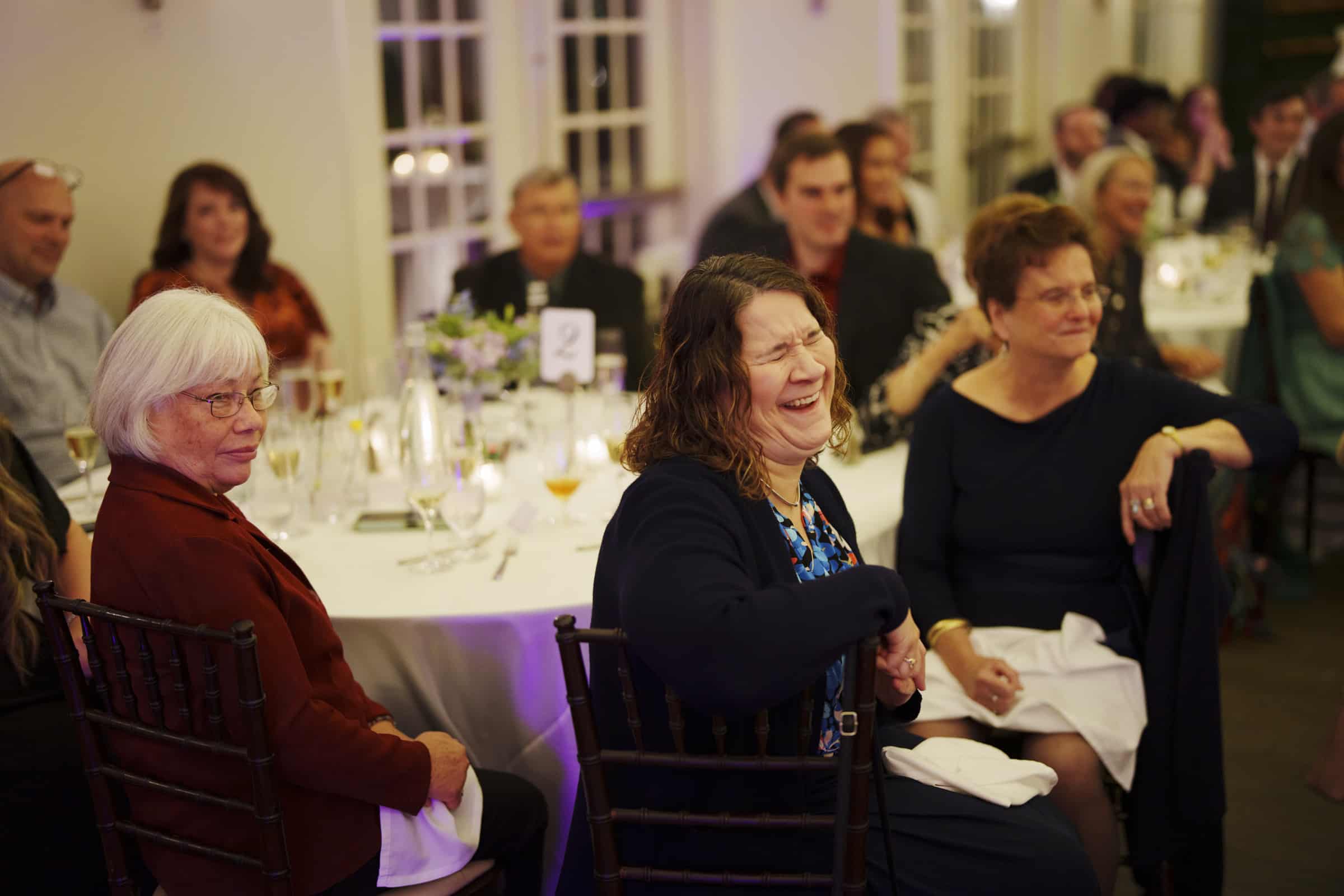Guests seated at round tables smile and laugh during a formal indoor event, such as a wedding reception. Glasses and floral centerpieces are on the tables, and people are dressed in semi-formal attire.