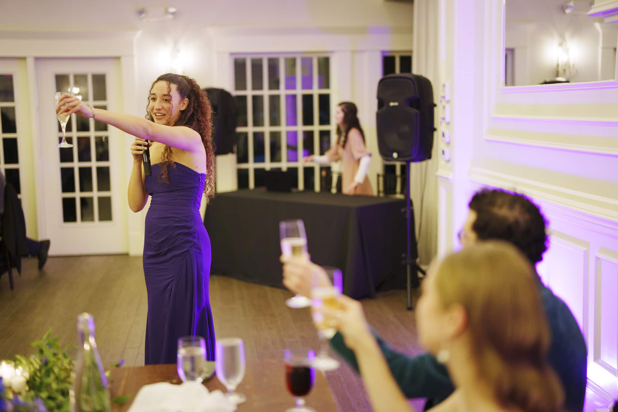 A woman in a purple dress gives a toast with a microphone, smiling and raising a glass. Two seated guests in the foreground also raise their glasses. The setting appears to be a formal indoor event or celebration.