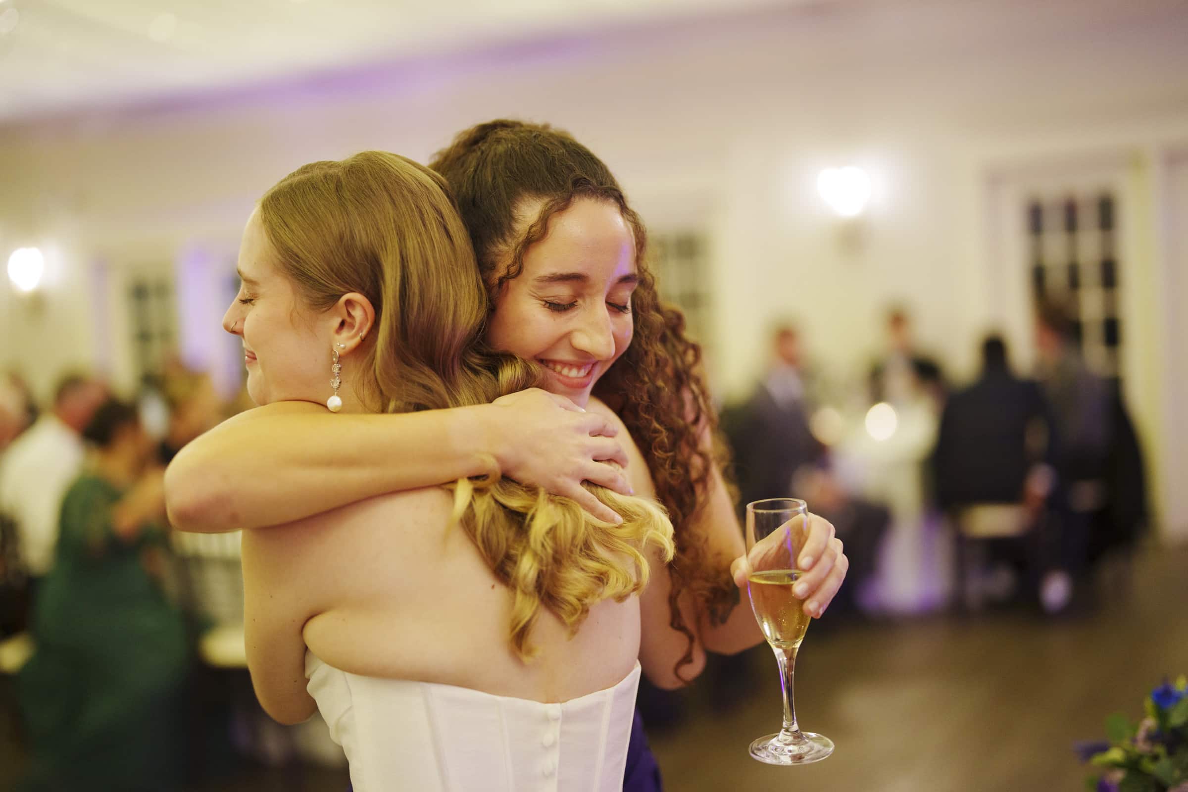 Two women embrace and smile warmly at a formal event; one holds a glass of champagne. The background shows people seated at tables, suggesting a celebration or wedding reception.