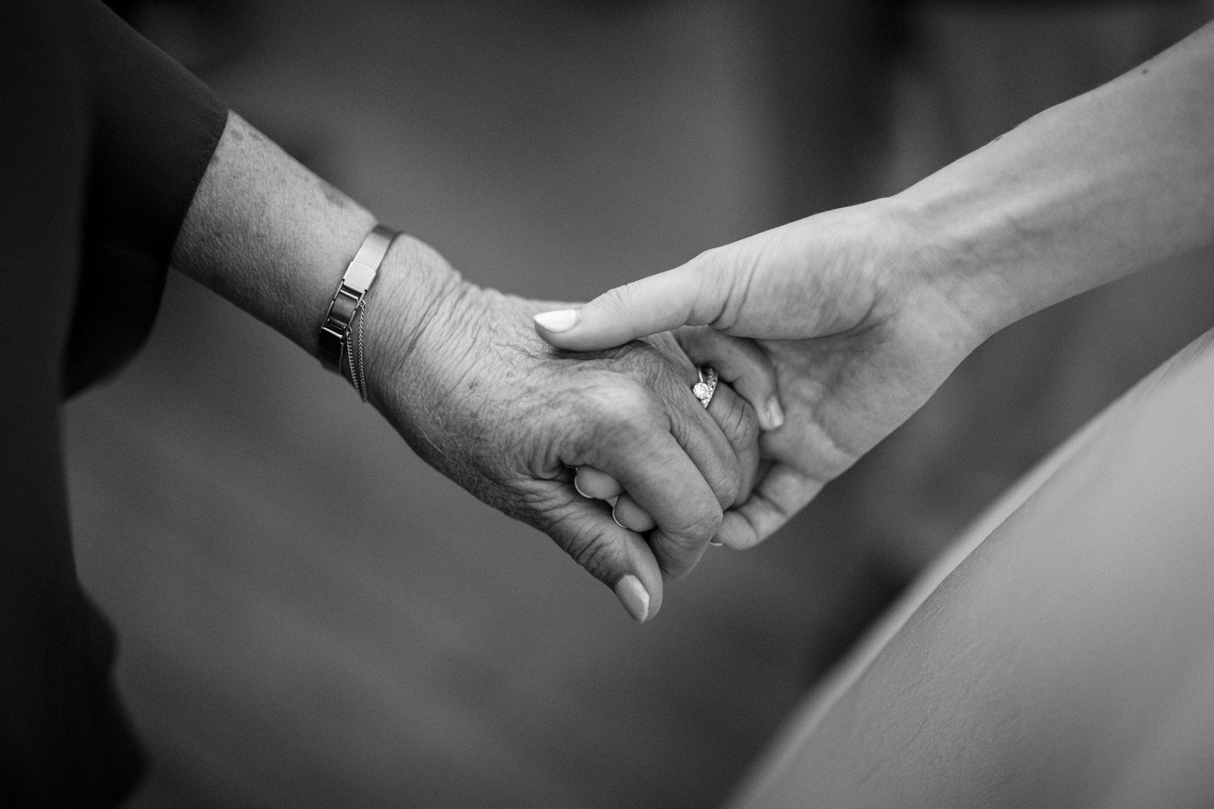 Black and white photo of two people holding hands—one older with a watch and one younger with an engagement ring. The image conveys tenderness and connection across generations.