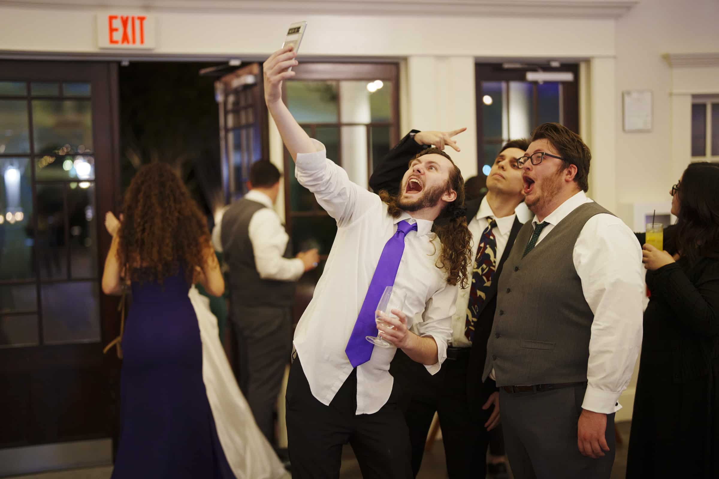 A group of men dressed in formal attire pose energetically for a selfie at an indoor event, one holding up a phone while others make playful expressions. People mingle in the background near open double doors.