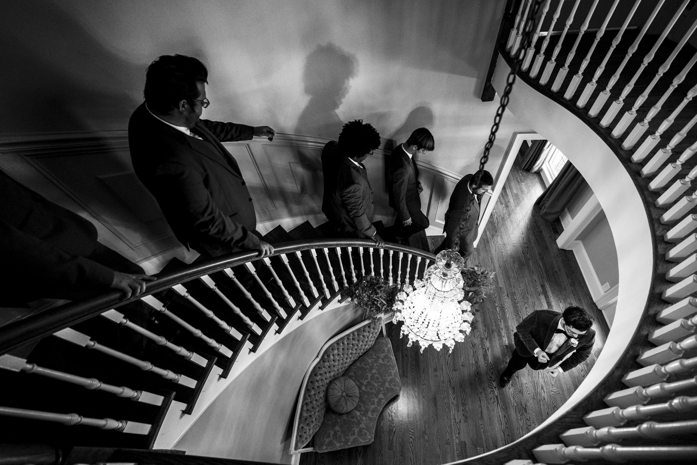 A black-and-white photo shows five men in suits descending a curved staircase. One man stands below by a chandelier, looking at something in his hands. The scene is elegant and formal, with strong shadows.