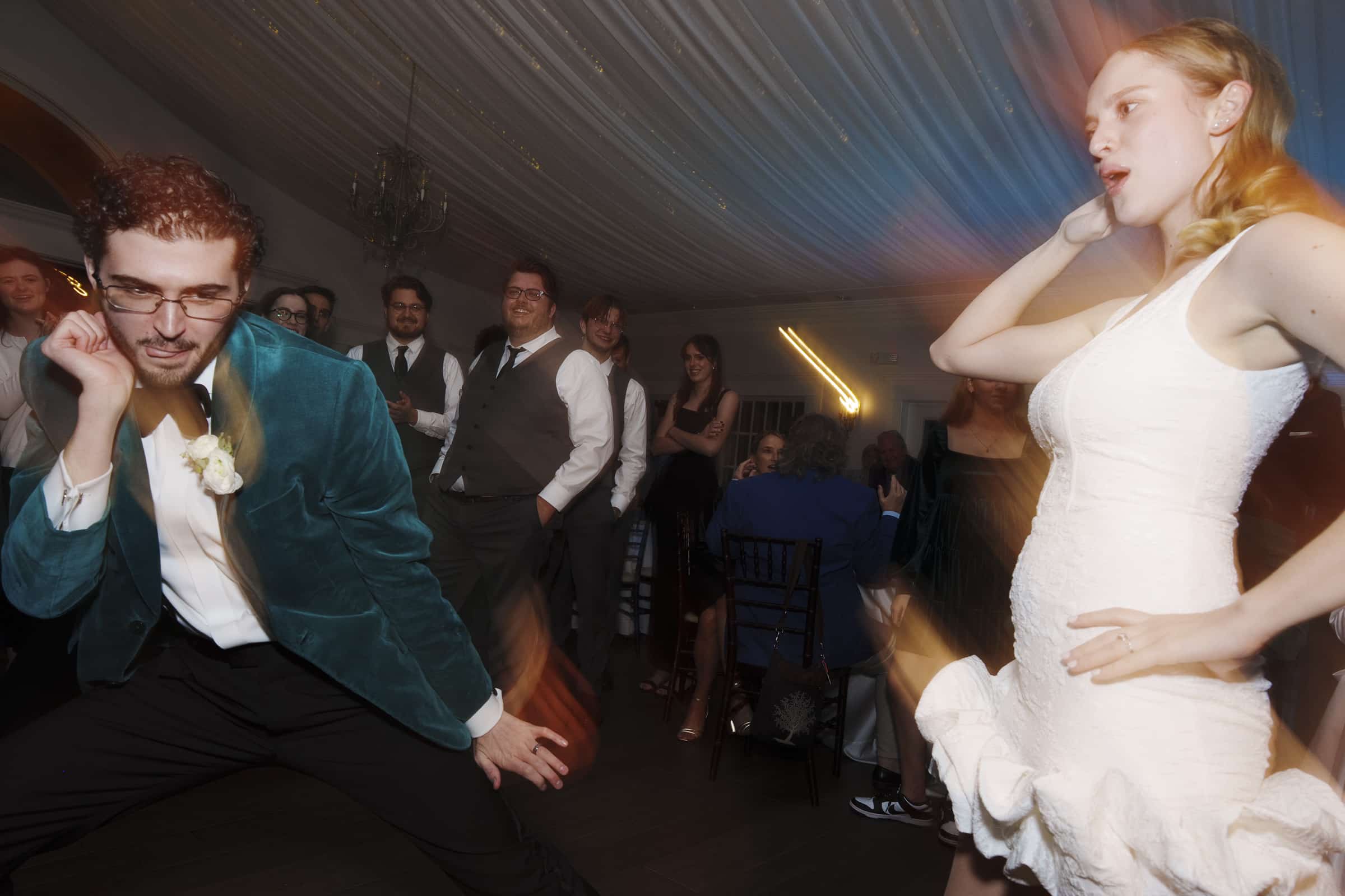 A man in a green velvet jacket and a woman in a white dress dance enthusiastically at a lively indoor event, surrounded by smiling and cheering guests under a draped ceiling.