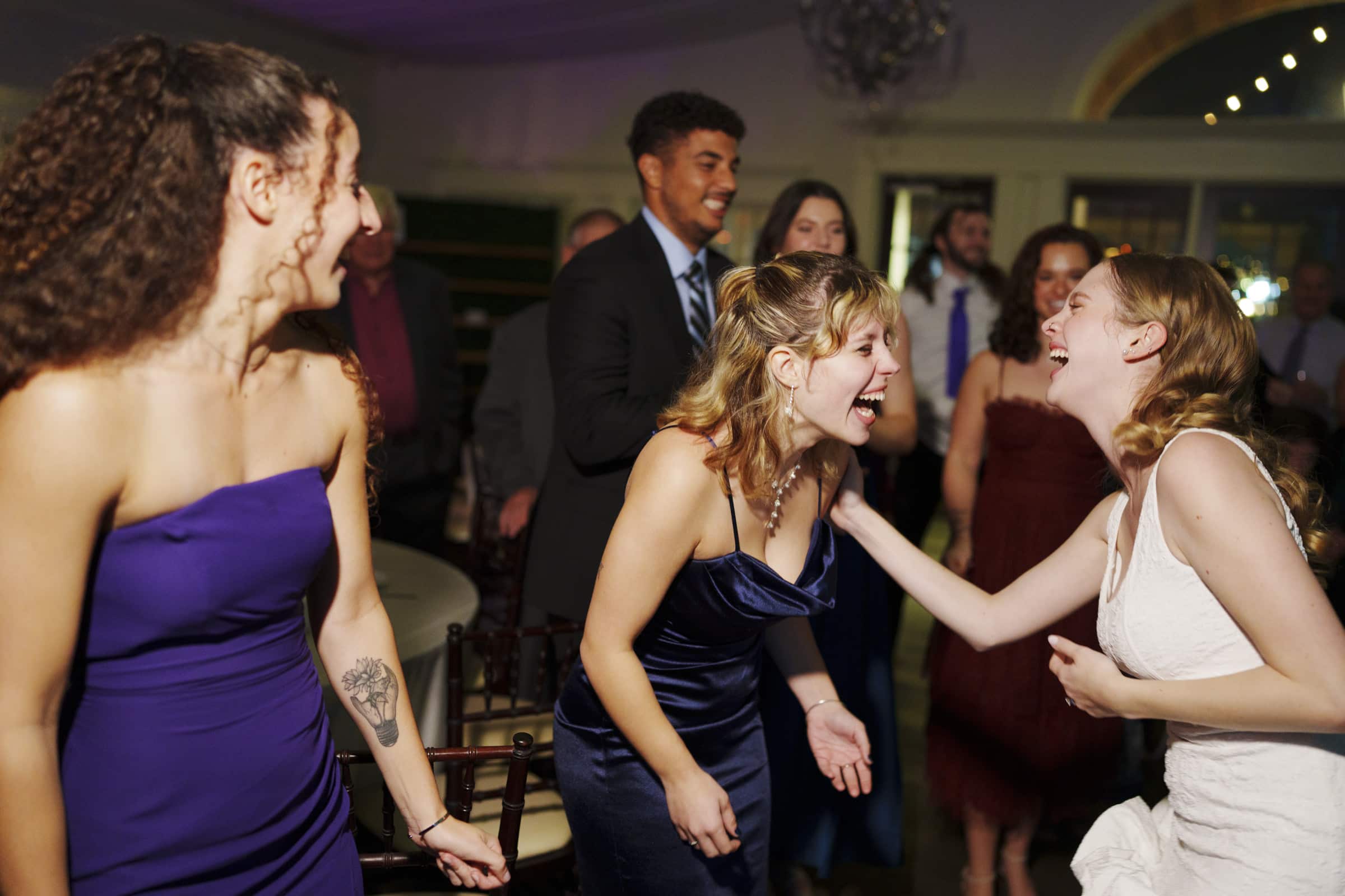 A group of people are laughing and dancing at a formal event. Three women in dresses are in the foreground, including one in a white dress who appears to be the bride. Other guests in suits and dresses are smiling in the background.