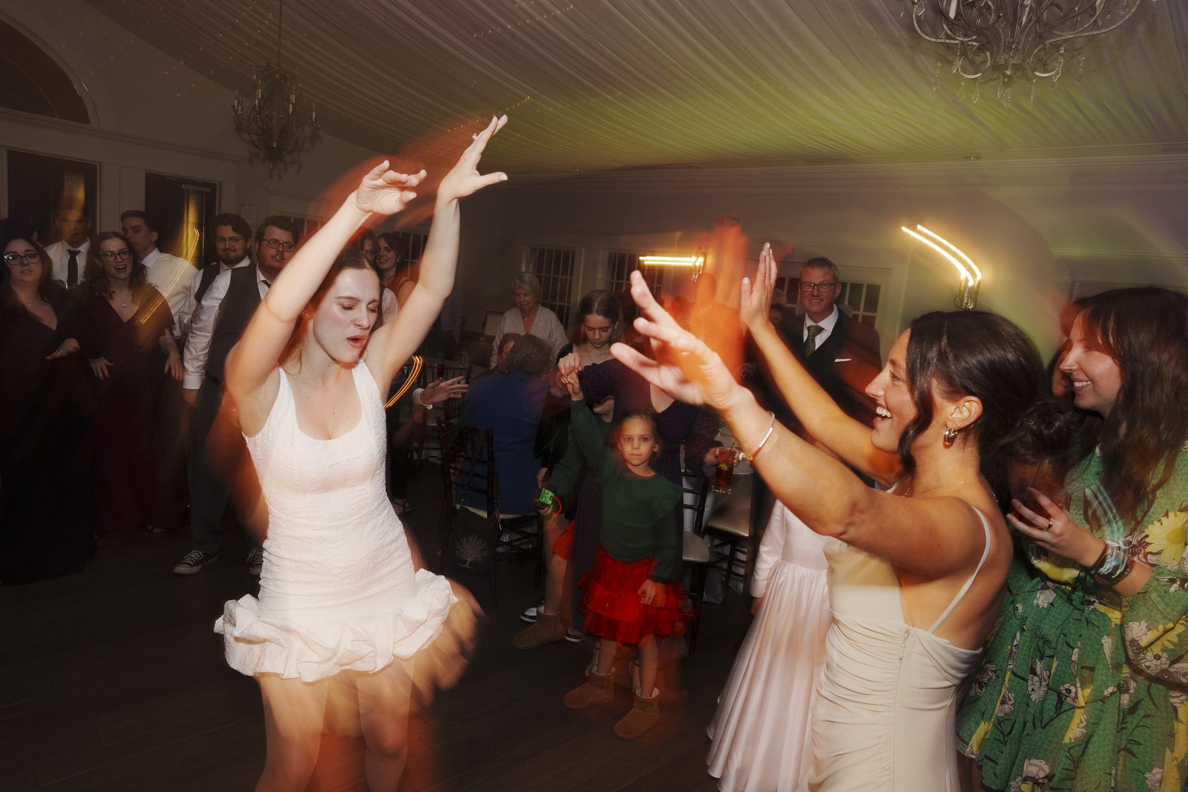 Two women dance energetically with raised arms at a lively indoor party, surrounded by smiling guests. Some people clap, and children watch. Warm, colorful lights blur with motion, capturing the joyful atmosphere.