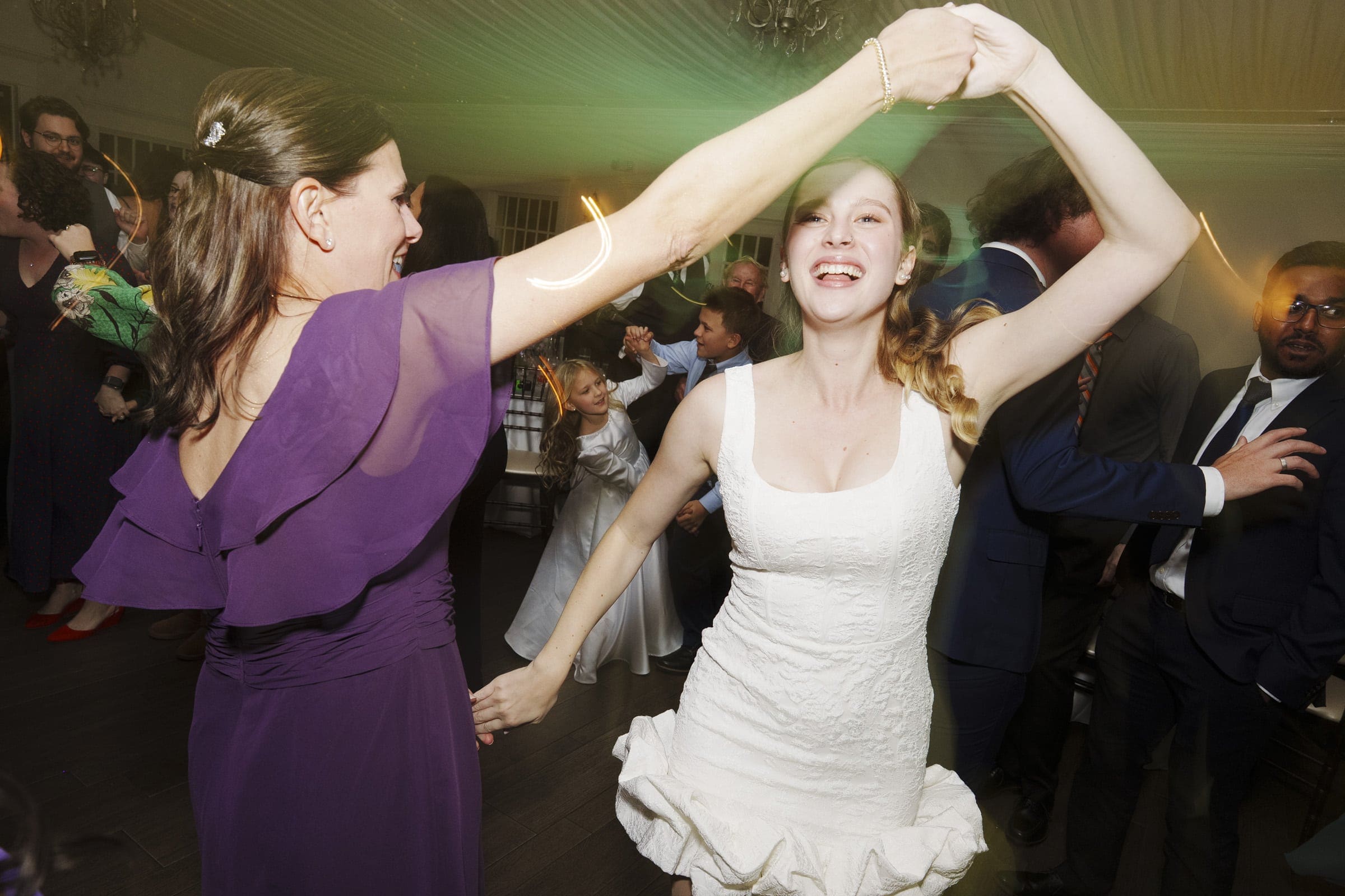 Two women, one in a white dress and the other in a purple dress, joyfully dance together at a lively party surrounded by other guests. The atmosphere is festive, with motion and colorful lights.