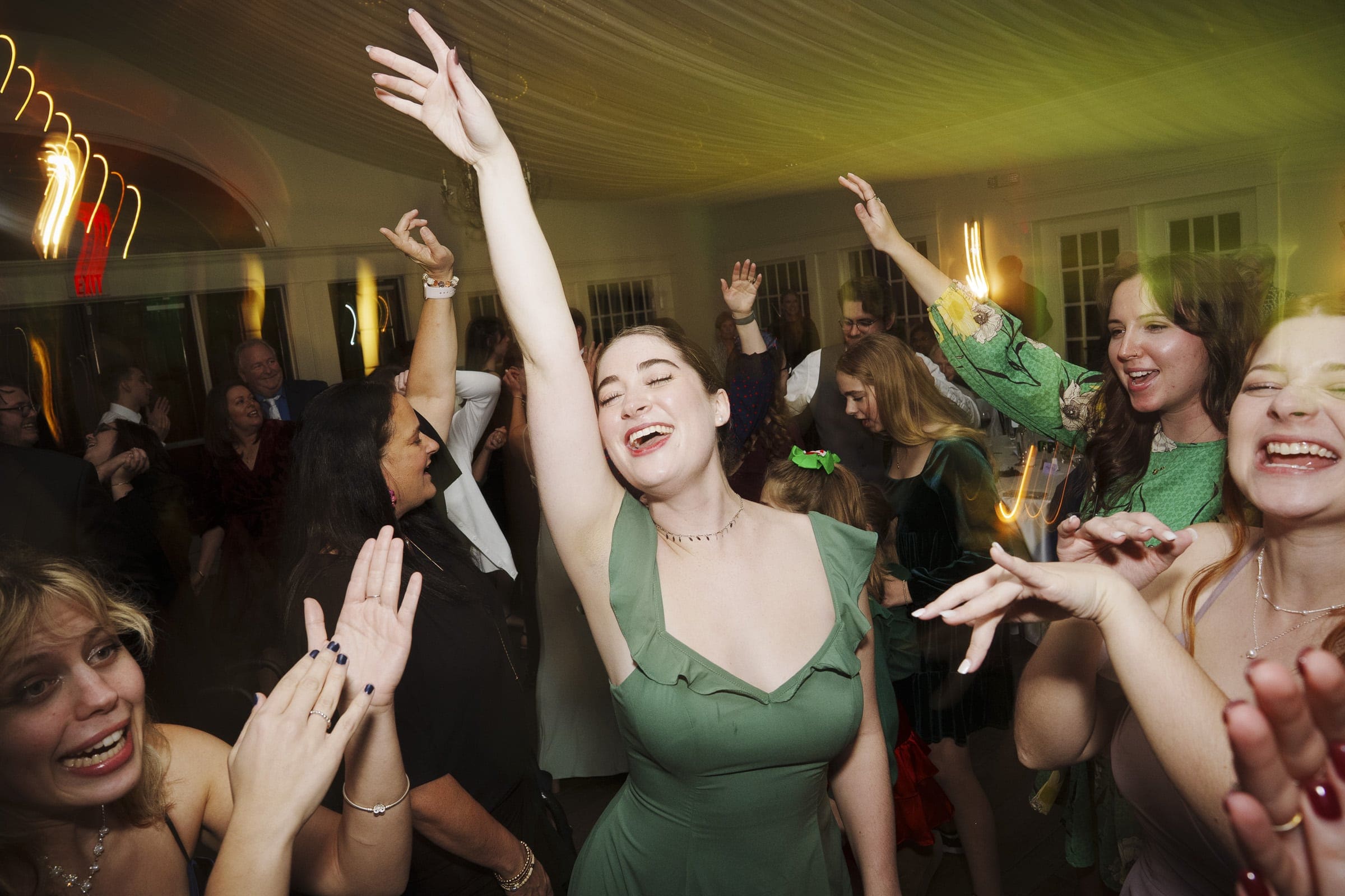A group of people in formal attire joyfully dance and celebrate at an indoor event, with one woman in a green dress raising her arm enthusiastically, surrounded by smiling and clapping guests.