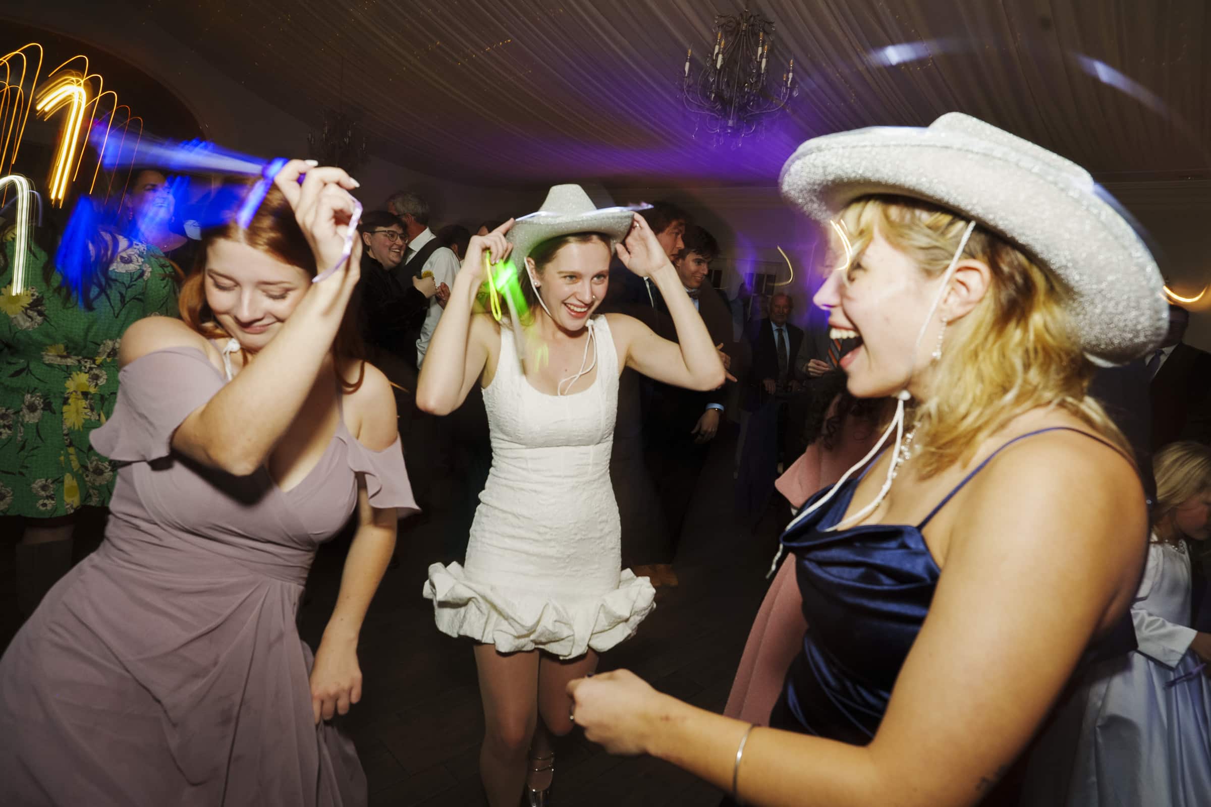 Three women in dresses and cowboy hats dance and laugh at a lively indoor party, with colorful lights and other people celebrating in the background.