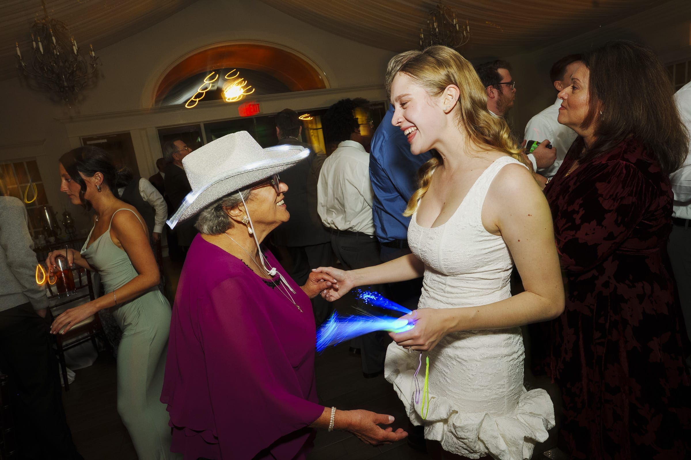 An elderly woman in a white cowboy hat and purple outfit smiles and holds hands with a young woman in a white dress at a lively indoor party, with other guests mingling in the background.