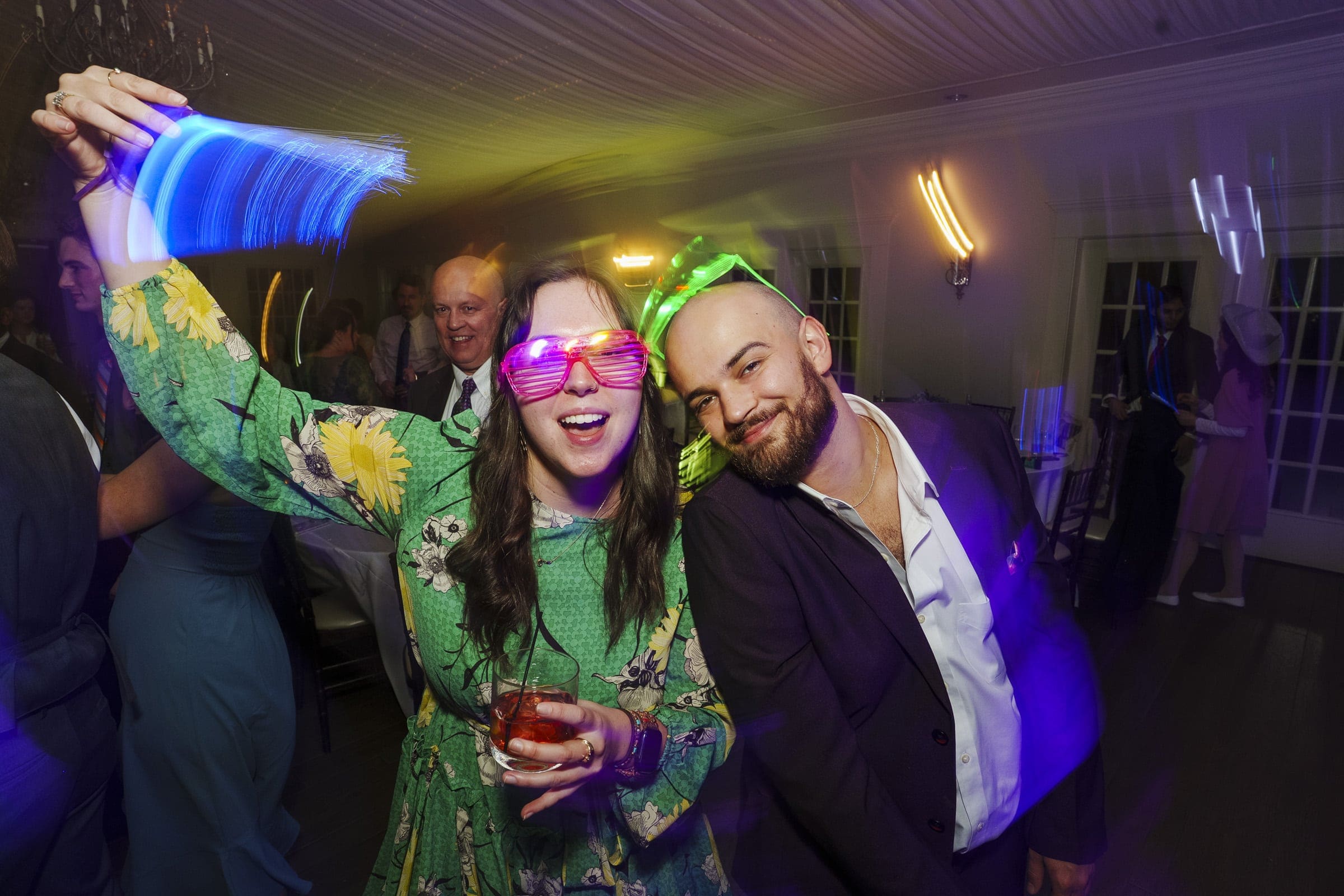 A woman in a green floral dress wears glowing pink shutter shades and waves a blue light stick while holding a drink. A smiling man in a suit leans in beside her at a lively indoor party.