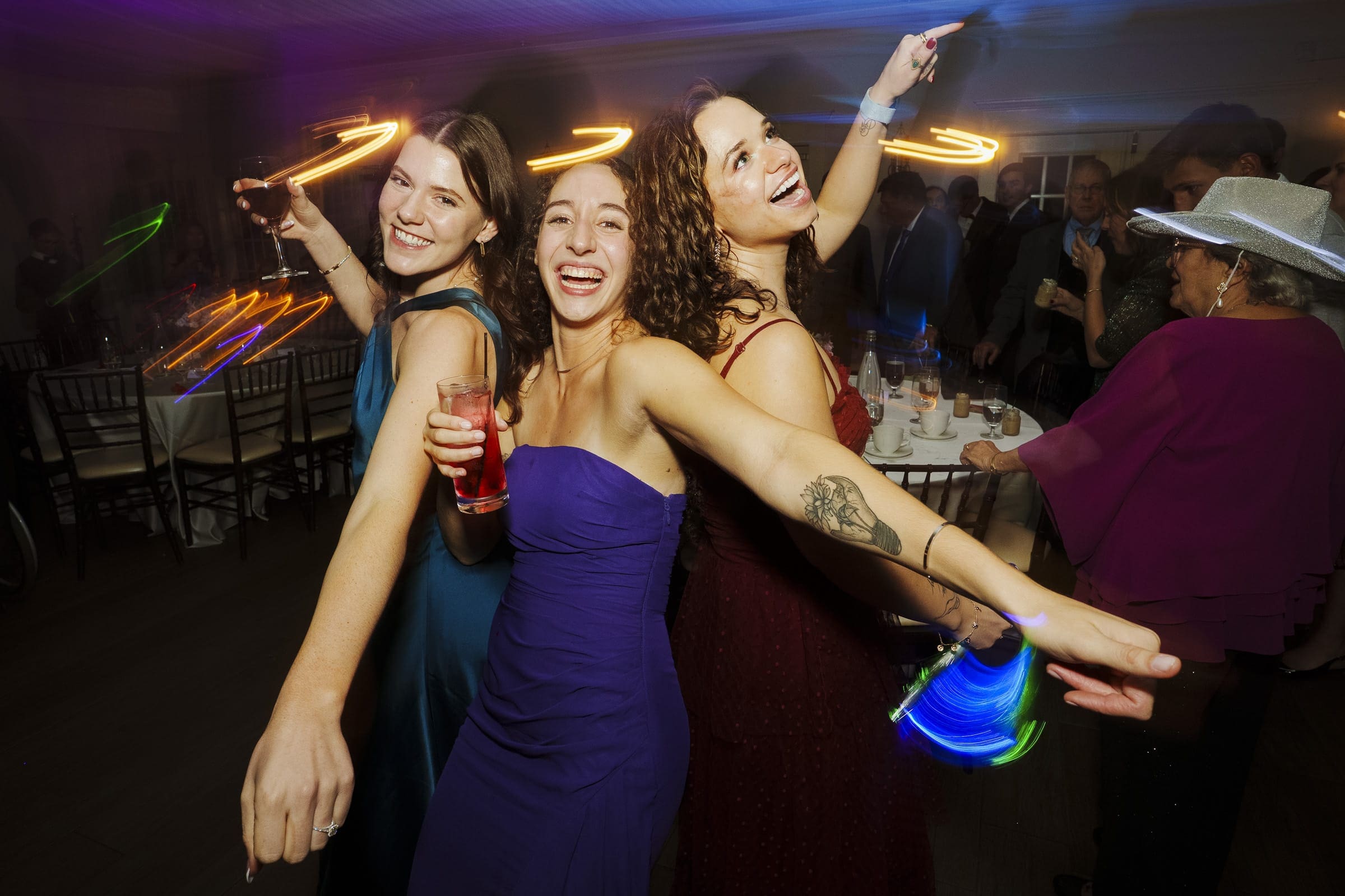 Three women in colorful dresses dance and smile at a lively indoor party with light trails in their hands, tables and guests visible in the background.