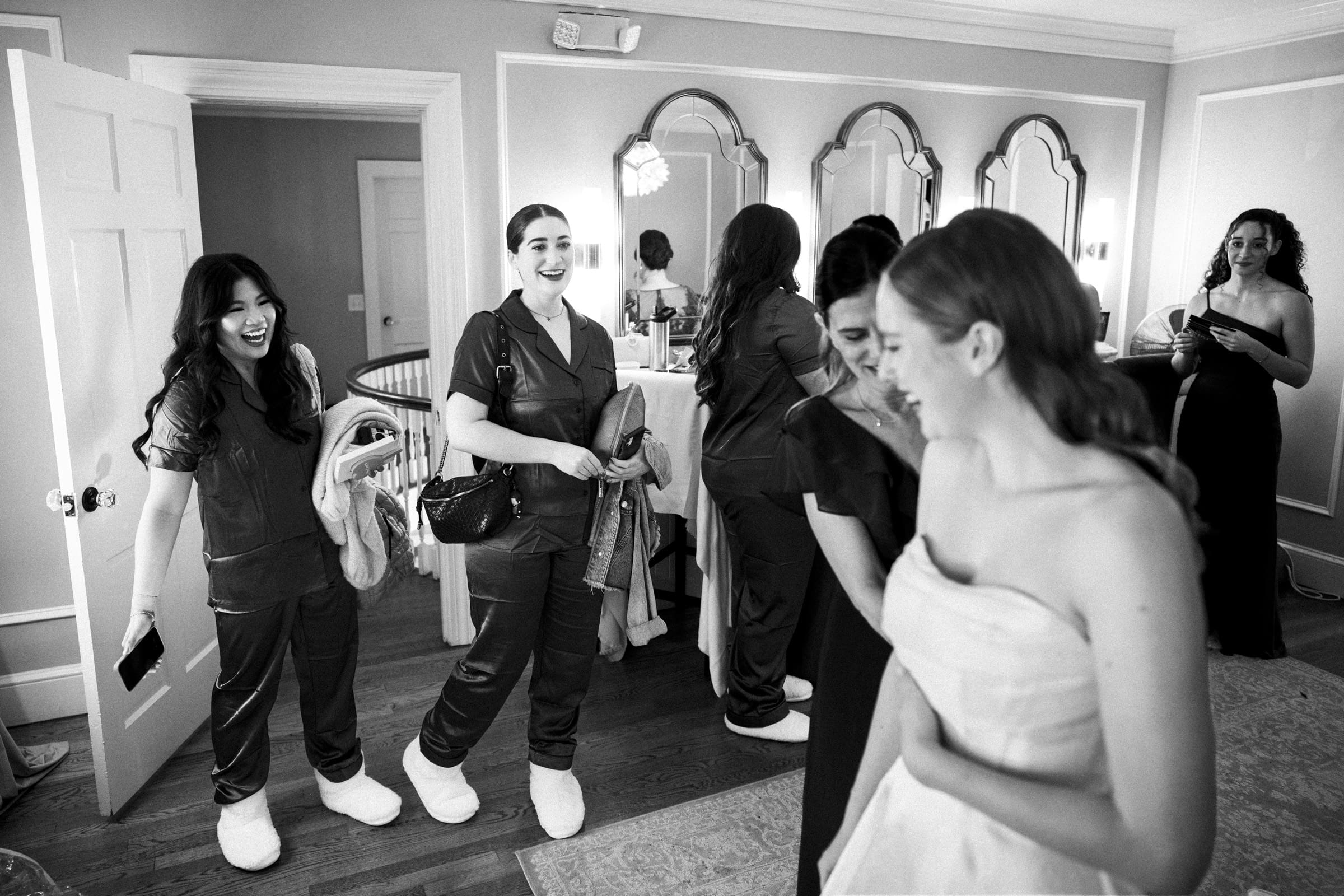 A bride smiles in a white dress as bridesmaids in matching pajamas and slippers laugh and chat in a bright, elegant room with mirrors and wooden floors.