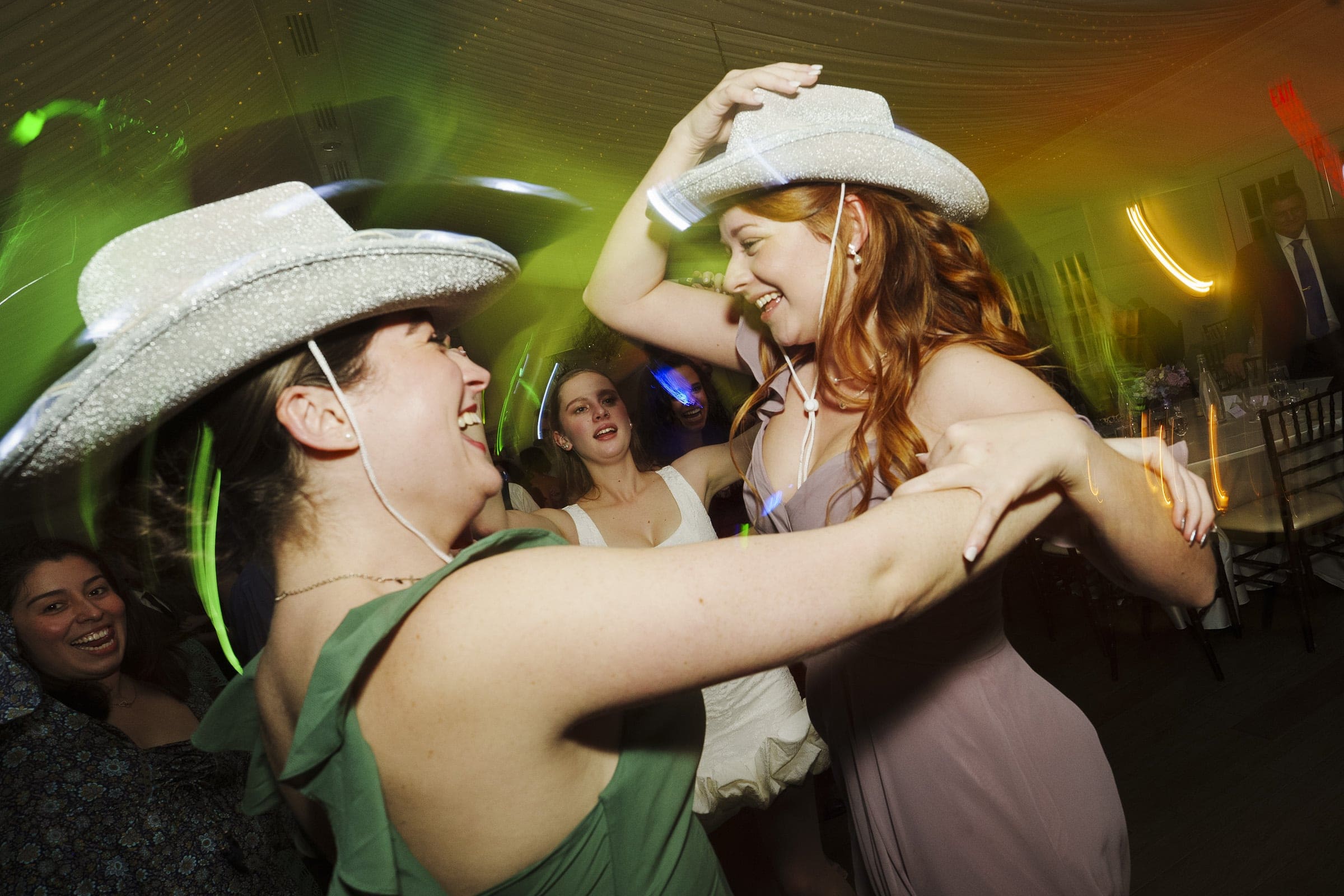 Two women wearing sparkly cowboy hats dance joyfully together at a lively indoor event, smiling and holding hands, with other people and colorful lights in the background.