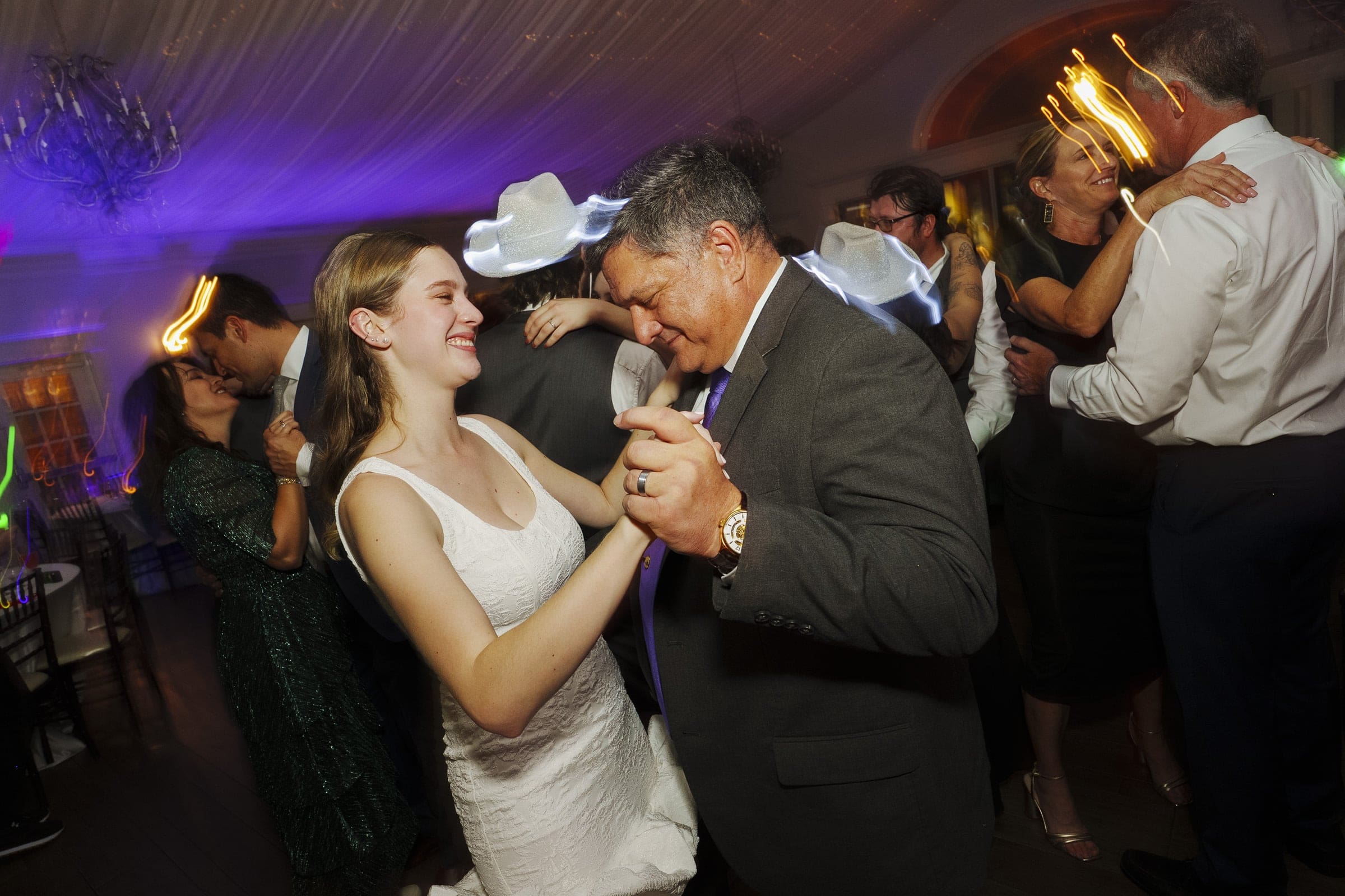 A young woman in a white dress and a man in a suit smile and dance together at a lively indoor celebration, surrounded by other couples dancing under colorful, festive lighting.