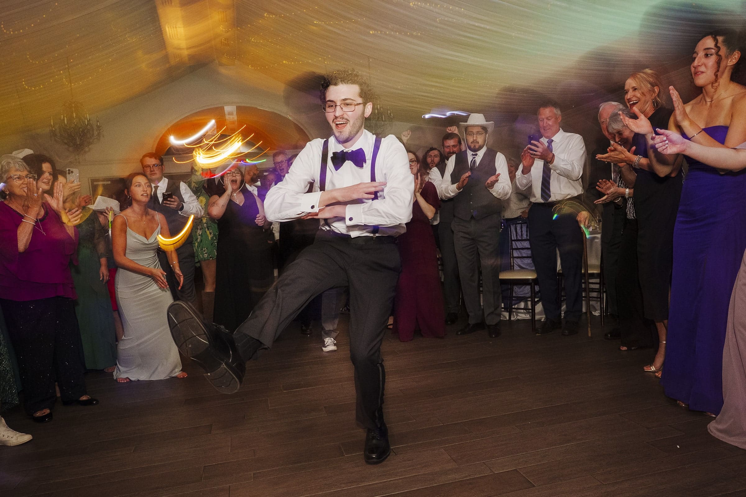 A man in formal attire dances energetically in the center of a lively crowd at an indoor event, while people around him clap, smile, and cheer him on. Warm lights add a festive, celebratory atmosphere.