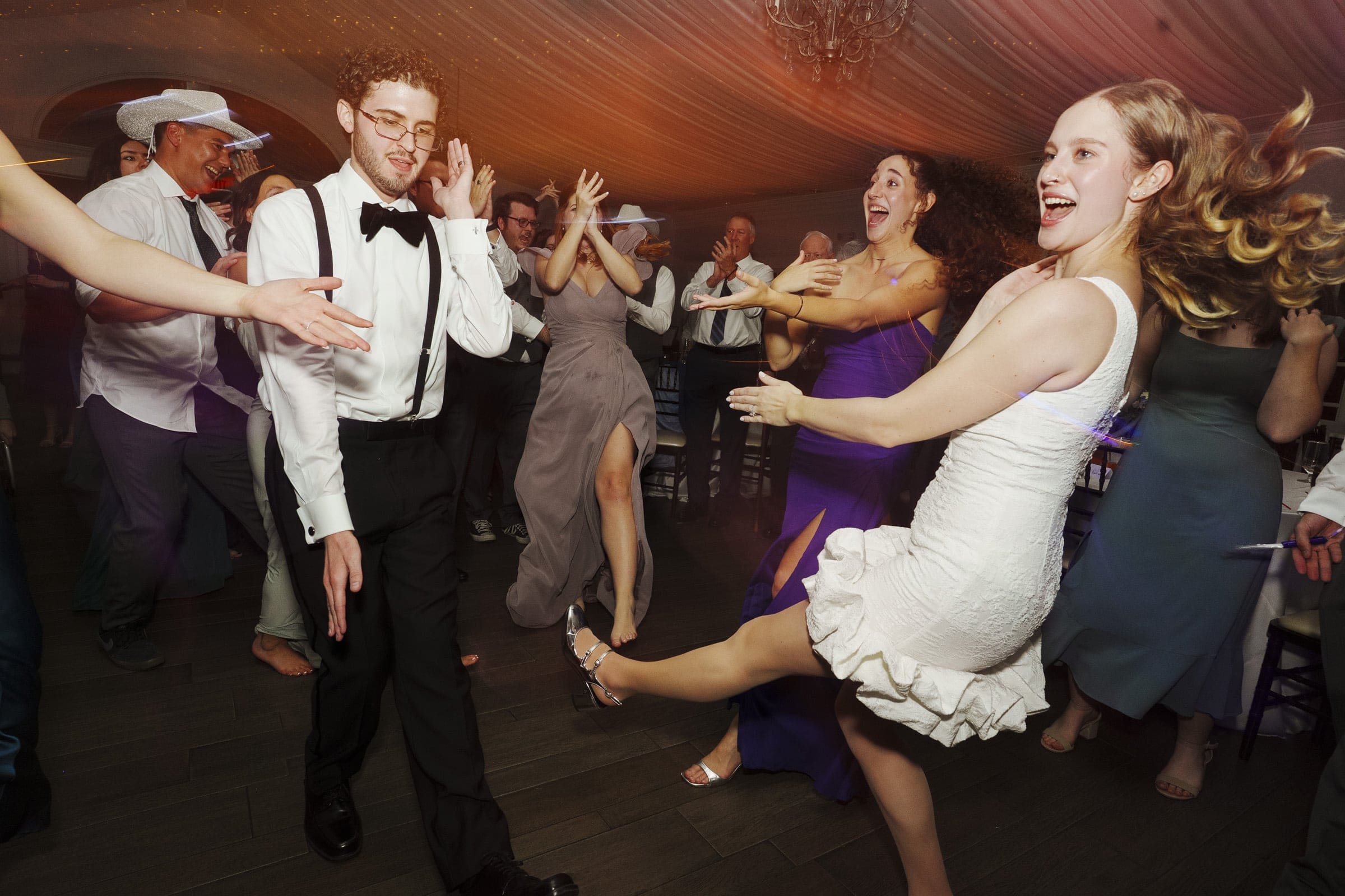 A group of people in formal attire dance energetically at a lively party or wedding reception, with joyful expressions and dynamic movement visible on the dance floor.