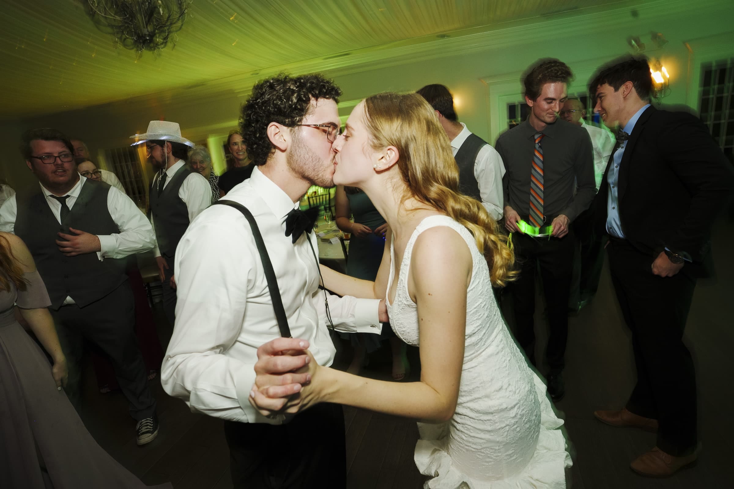 A bride and groom kiss while dancing at their wedding reception, surrounded by guests dressed in formal attire, some smiling and watching them. The room is warmly lit with green and yellow lights.
