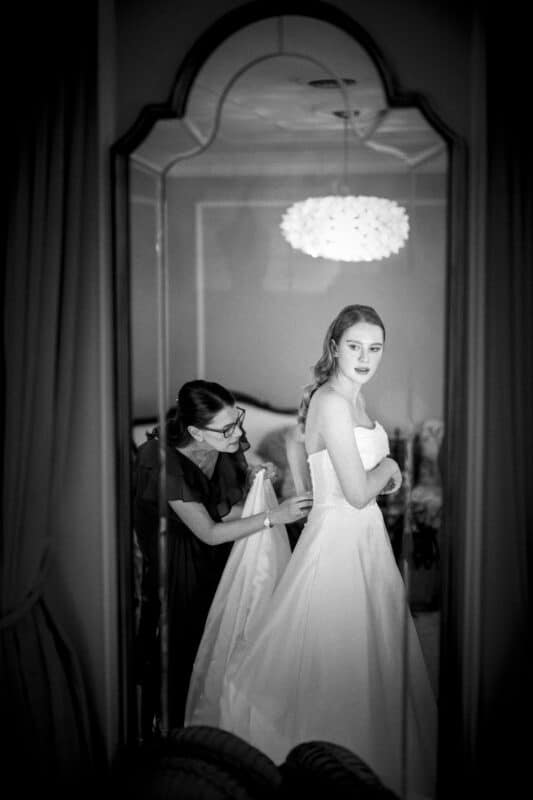 Black and white photo of a woman in a wedding dress being helped by another woman, reflected in an ornate mirror. Elegant room with soft lighting and a decorative ceiling light above.