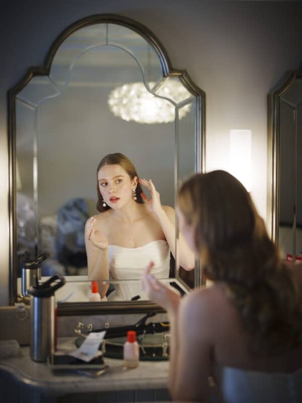 A woman in a white strapless dress puts on earrings while looking at herself in a mirror above a cluttered vanity. The scene is softly lit, with her reflection in focus and her figure blurred in the foreground.