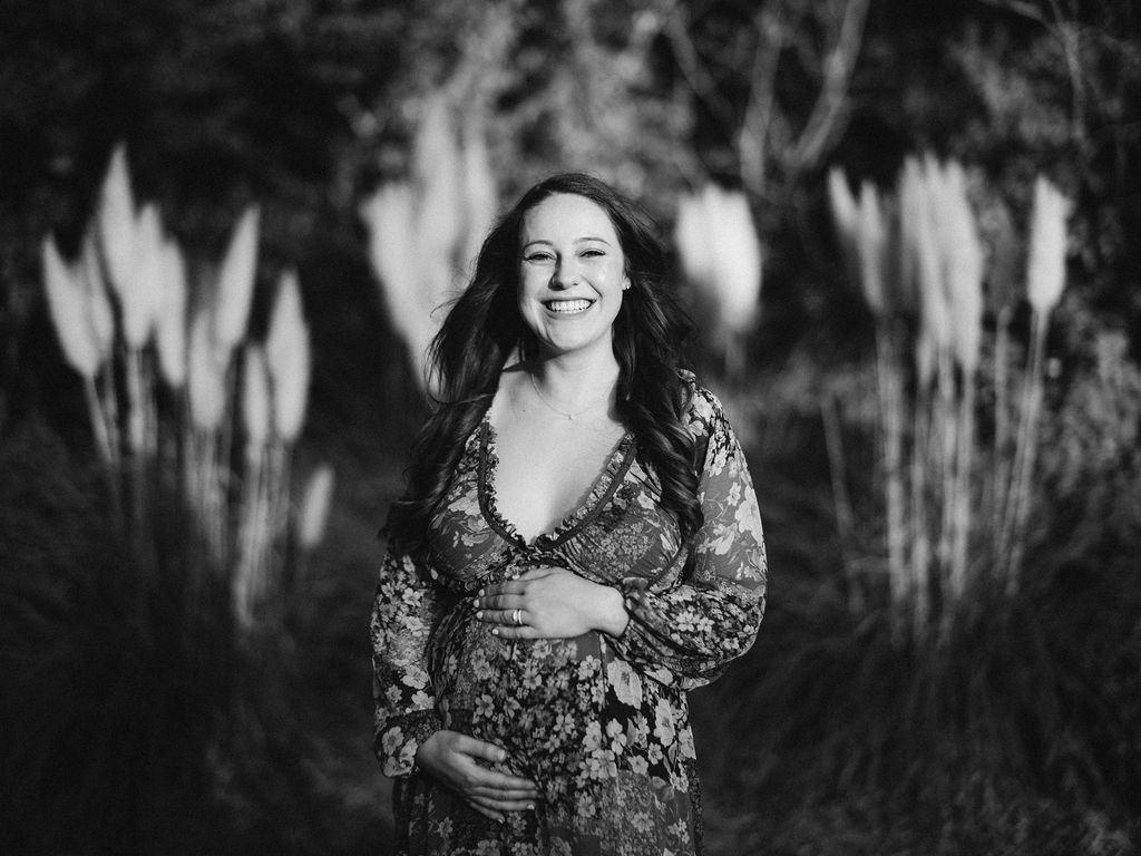 A smiling pregnant woman in a floral dress stands outdoors, holding her belly with both hands during one of our Autumn Mini Sessions. Tall grasses and blurred foliage fill the background. The photo is in black and white. By award-winning Raleigh wedding photographer, Joe Payne.