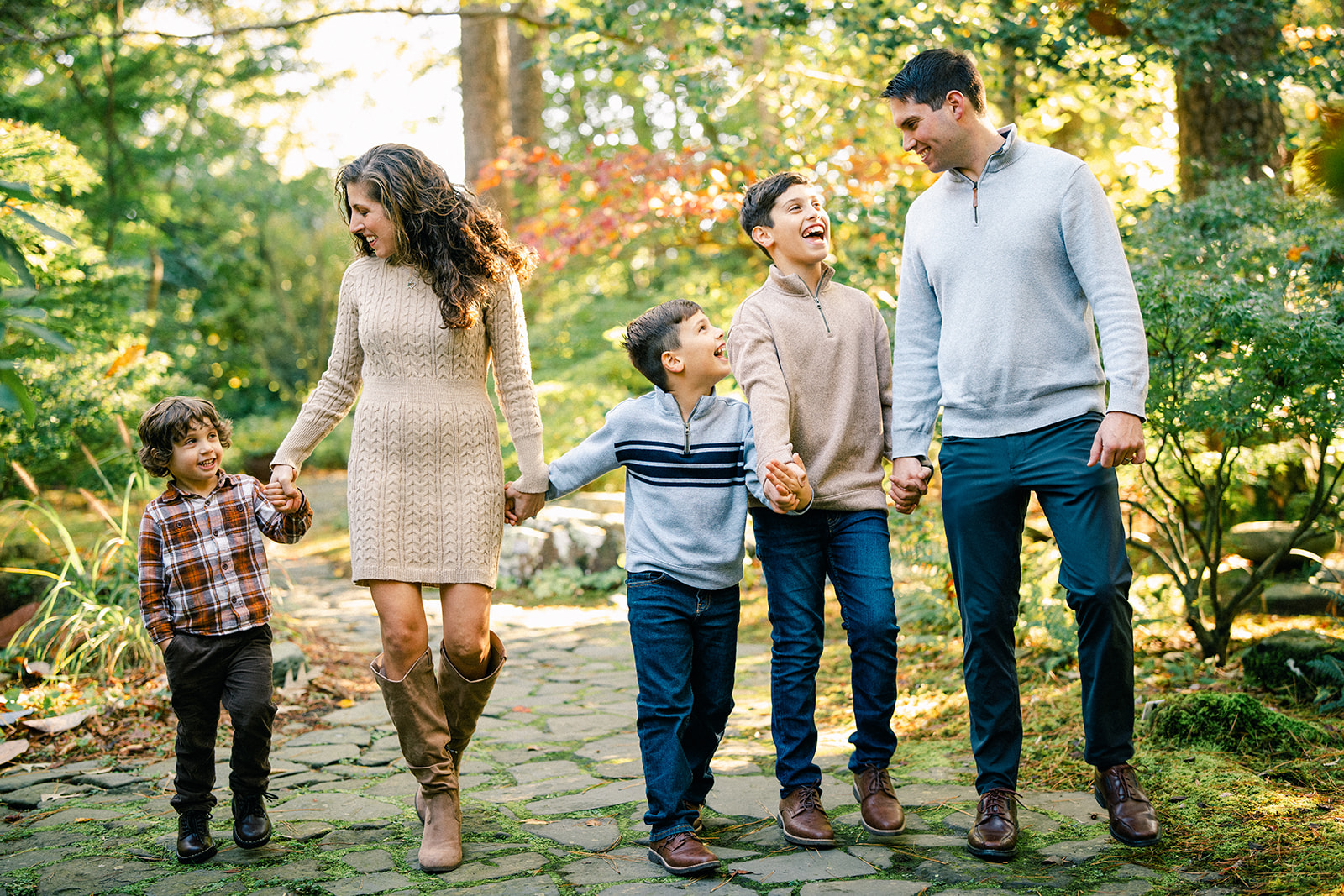 A family of five walks on a stone path in a garden, holding hands and smiling at each other during their Autumn Mini Session. The parents are on either end, with three children between them, as sunlight filters through the trees in the background. By award-winning Raleigh wedding photographer, Joe Payne.