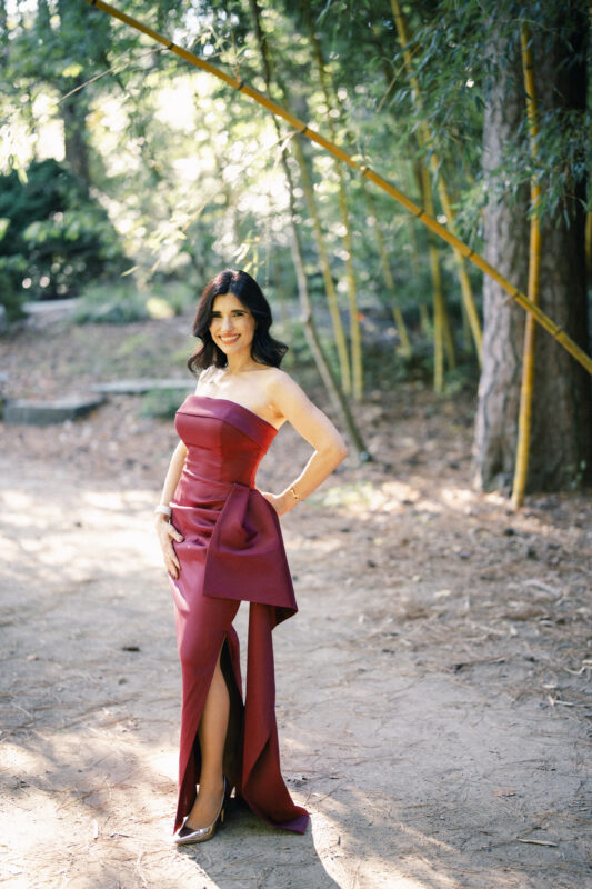 Woman in a strapless burgundy gown stands outdoors on a sunlit, wooded path surrounded by trees and bamboo, smiling with one hand on her hip—perfect inspiration for Autumn Mini Sessions. By award-winning Raleigh wedding photographer, Joe Payne.