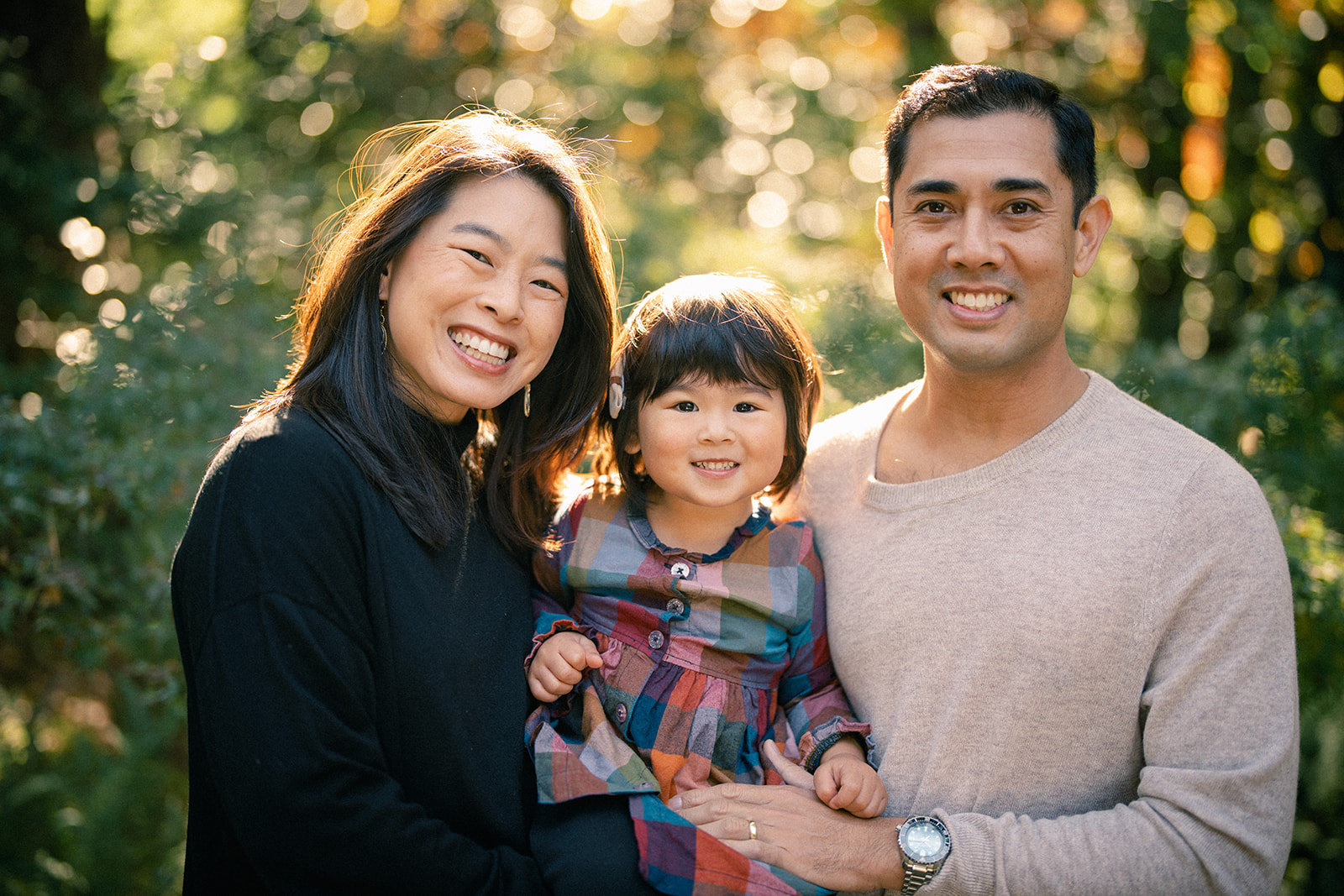 A smiling family of three stands outdoors in sunlight, with greenery in the background, capturing a perfect moment for Autumn Mini Sessions. The child, wearing a colorful dress, is held between two adults, who are both smiling warmly at the camera. By award-winning Raleigh wedding photographer, Joe Payne.