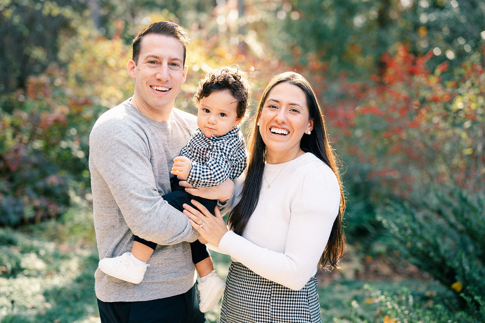 A man and woman smile at the camera outdoors, holding a young child during their Autumn Mini Session. The background is sunlit with greenery and colorful autumn leaves. By award-winning Raleigh wedding photographer, Joe Payne.