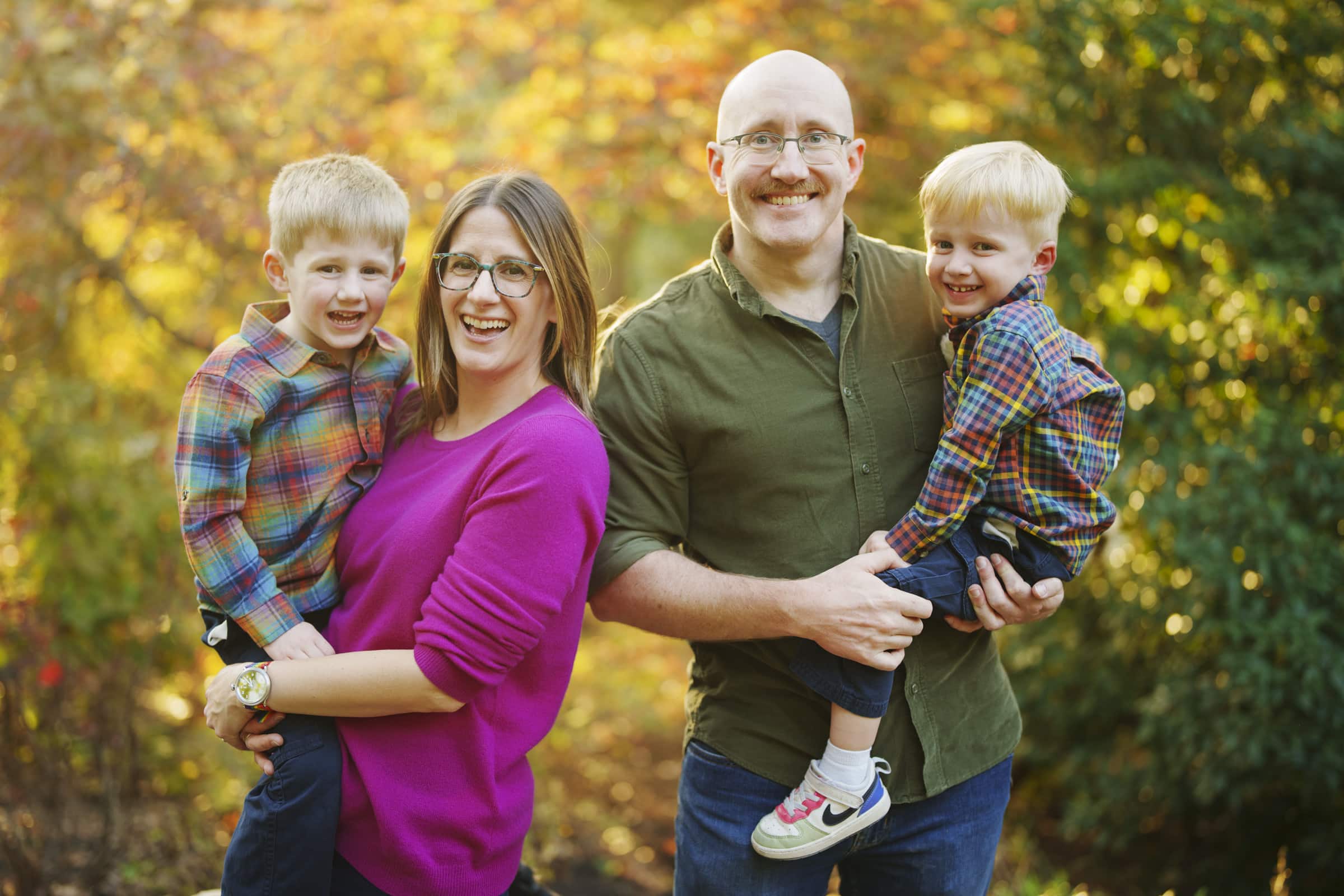 A smiling family of four poses outdoors in autumn. Two adults hold two young boys, all wearing colorful clothes. Trees with yellow and orange leaves fill the background.