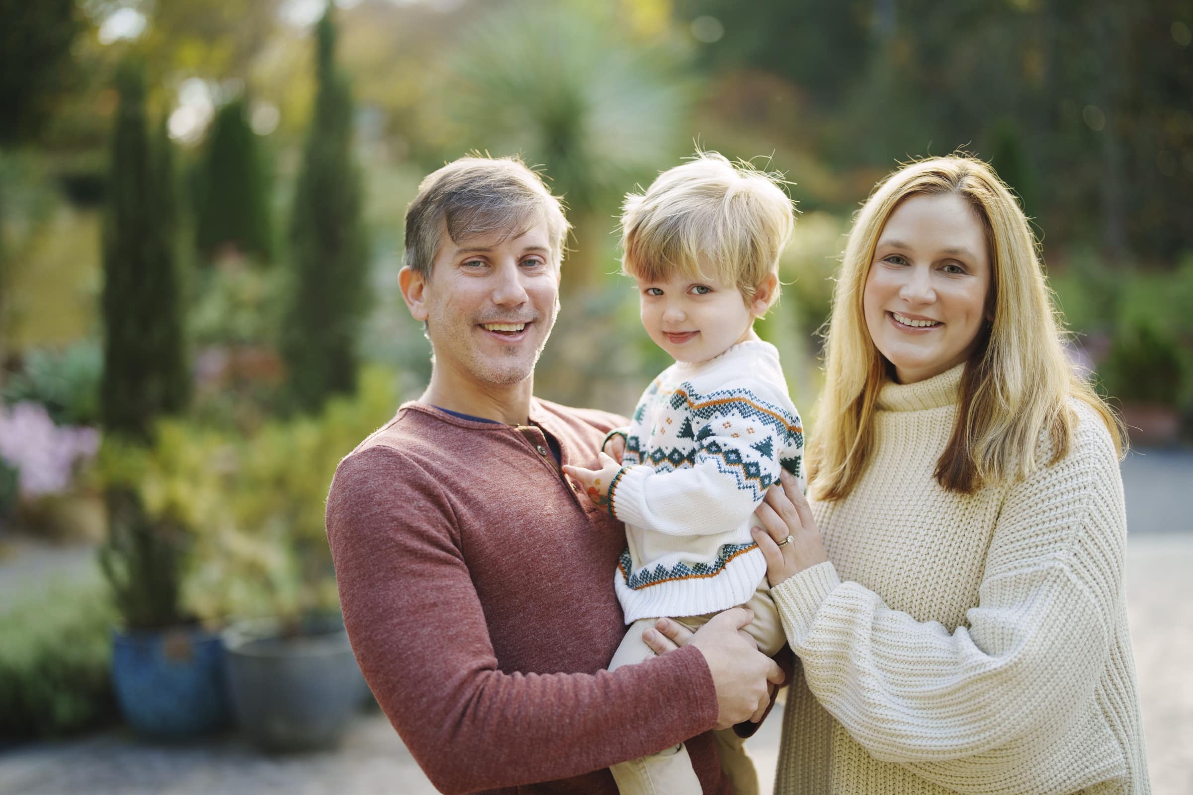 A smiling family of three stands outdoors. The father, in a red sweater, holds their young son, who wears a patterned sweater. The mother, in a cream turtleneck, stands beside them. Green plants and trees blur in the background.