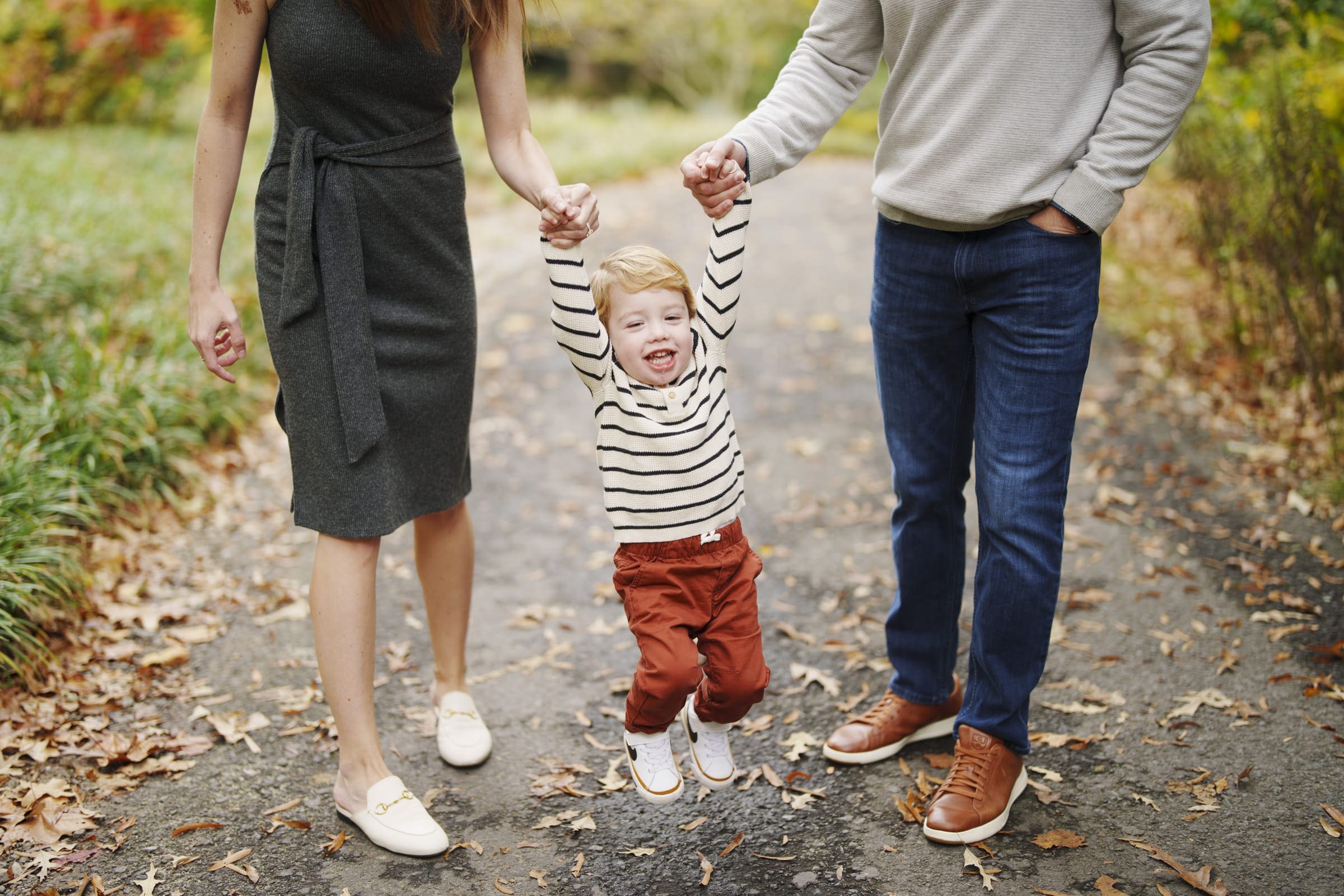 A young child in orange pants and a striped shirt is joyfully swinging between two adults holding his hands as they walk on a leaf-covered path outdoors. The adults’ faces are not visible.