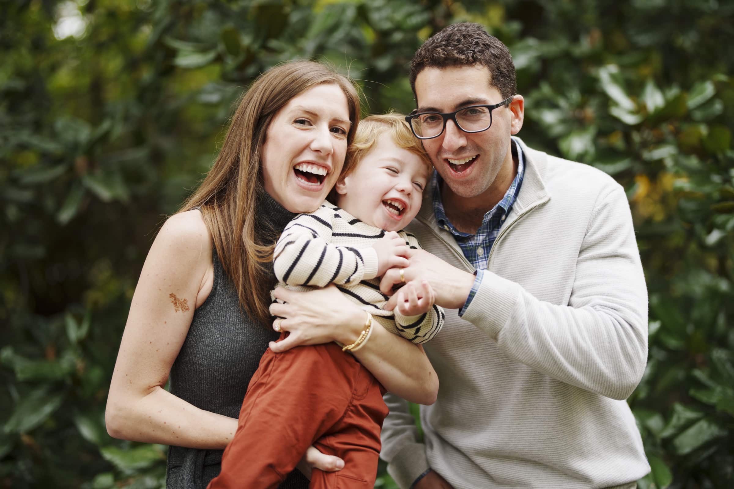 A smiling couple holds a laughing young child outdoors in front of green foliage, all appearing happy and enjoying a playful moment together.