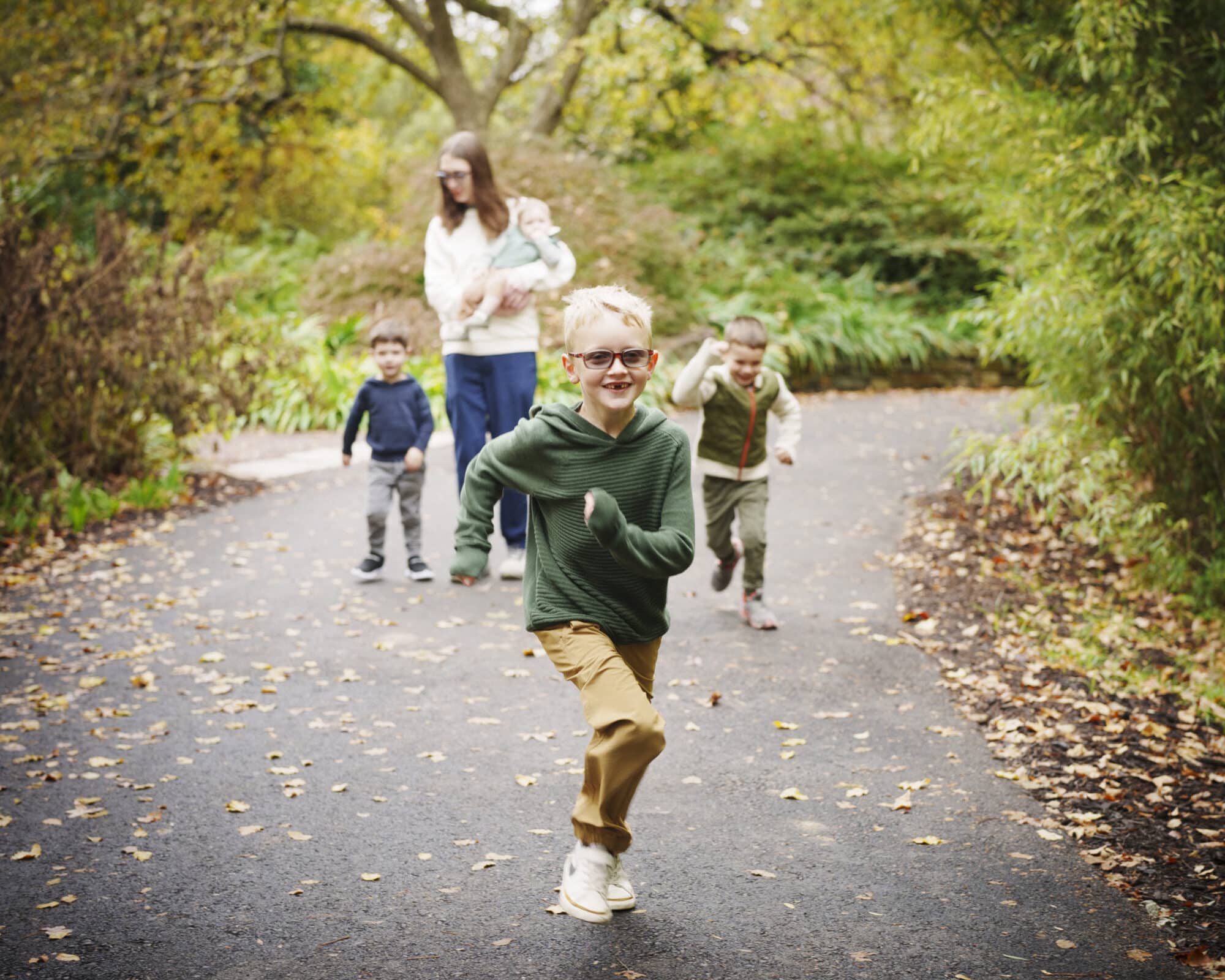 Three children run down a leaf-strewn path in a park, smiling and playing. Behind them, an adult carrying a baby walks along the path, surrounded by green trees and foliage.