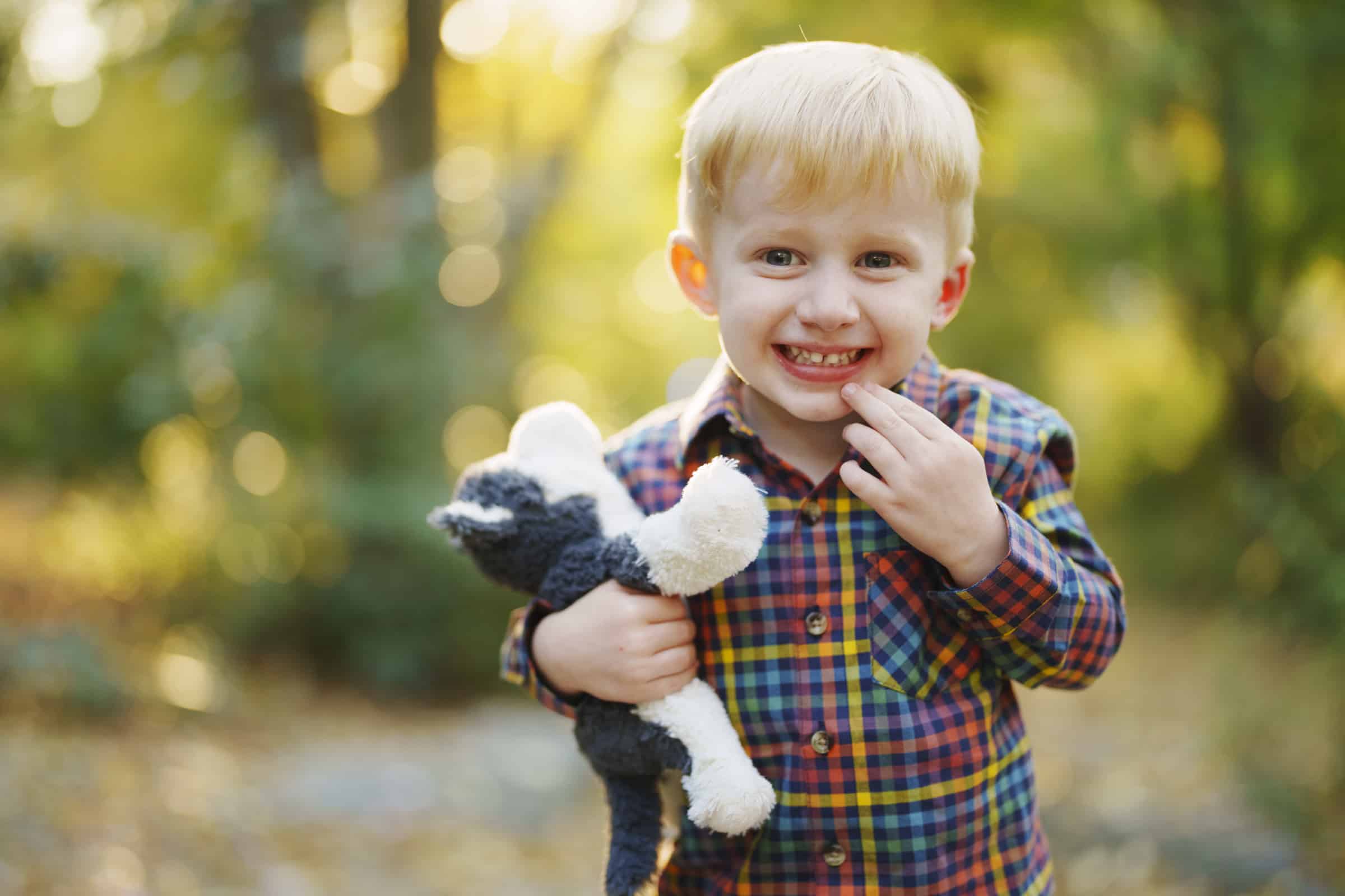 A young boy with blonde hair smiles while holding a stuffed animal in one hand and touching his mouth with the other, standing outdoors in a sunlit, leafy area.