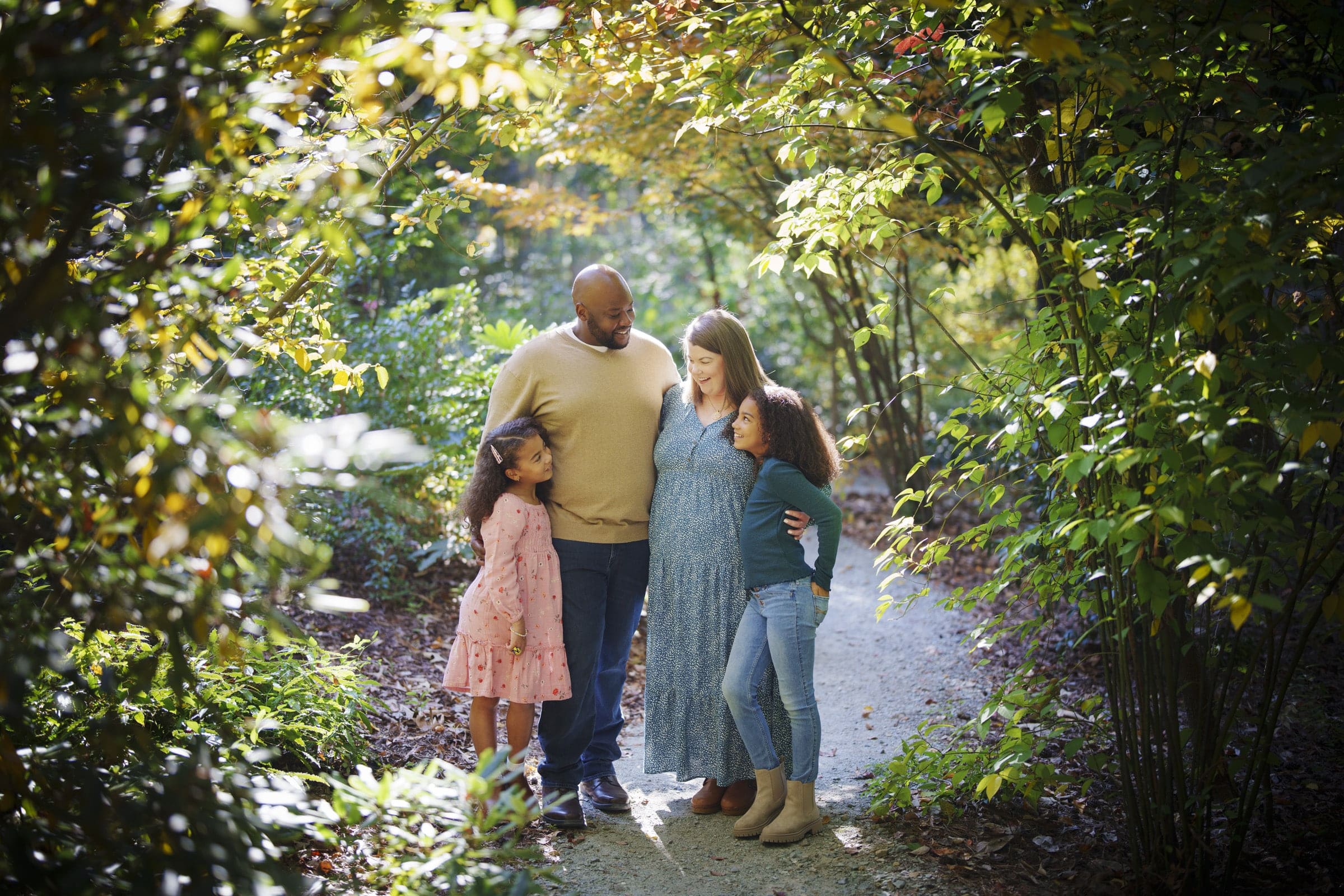 A family of four stands together on a sunlit forest path, surrounded by lush green trees. The parents smile at each other while their two young daughters, one in pink and one in blue, stand close by and look up at them.