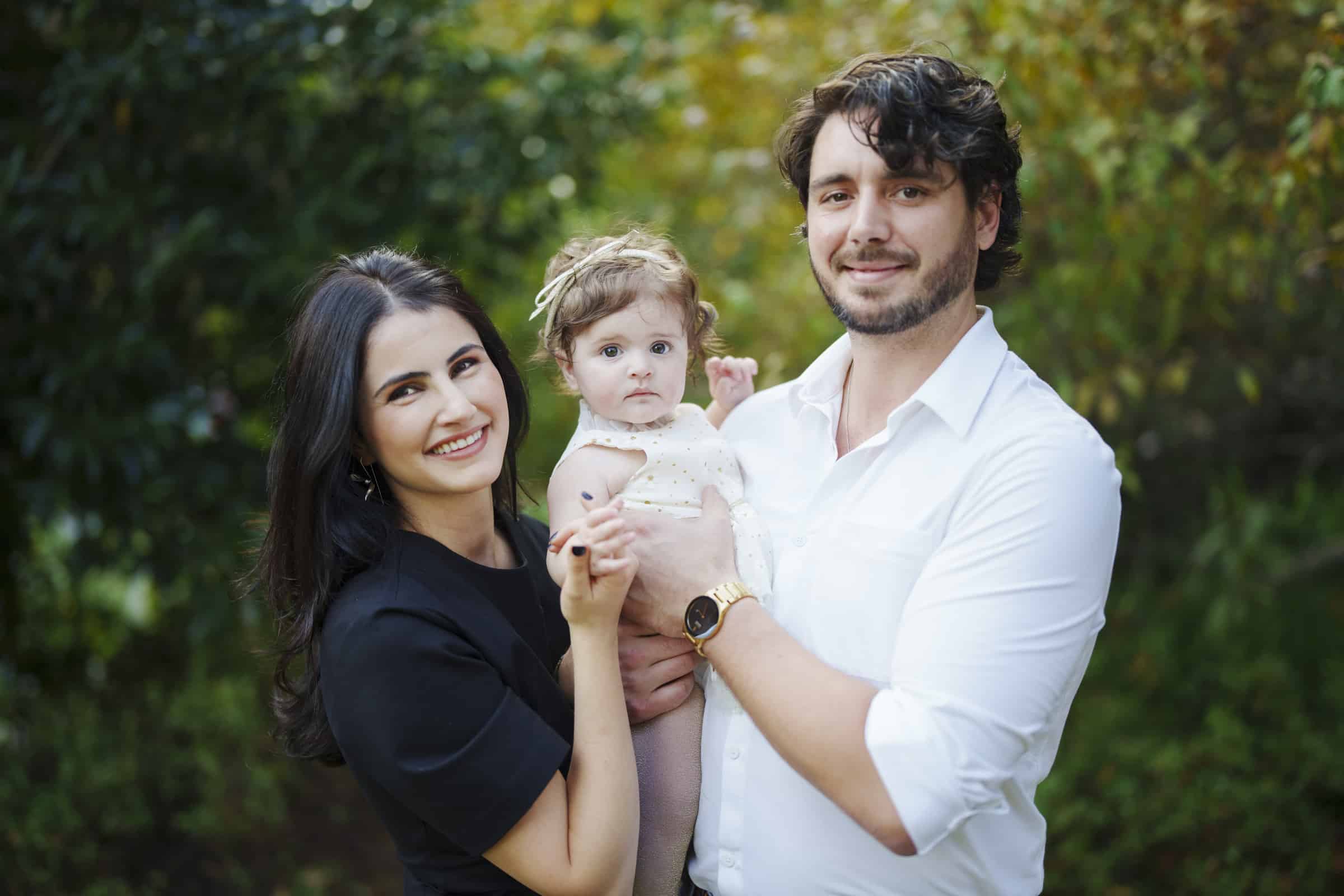 A smiling woman and man stand outdoors holding a baby between them. The woman has long dark hair and wears a black dress; the man has wavy hair, a beard, and a white shirt. The baby looks at the camera. Green foliage is in the background.