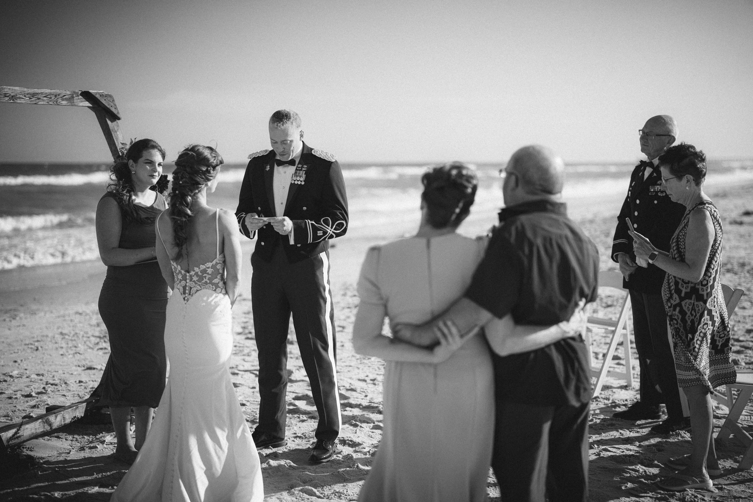 A small wedding ceremony takes place on a beach. The bride and groom stand facing each other, surrounded by a few guests, with the ocean in the background under clear skies. The scene is in black and white.