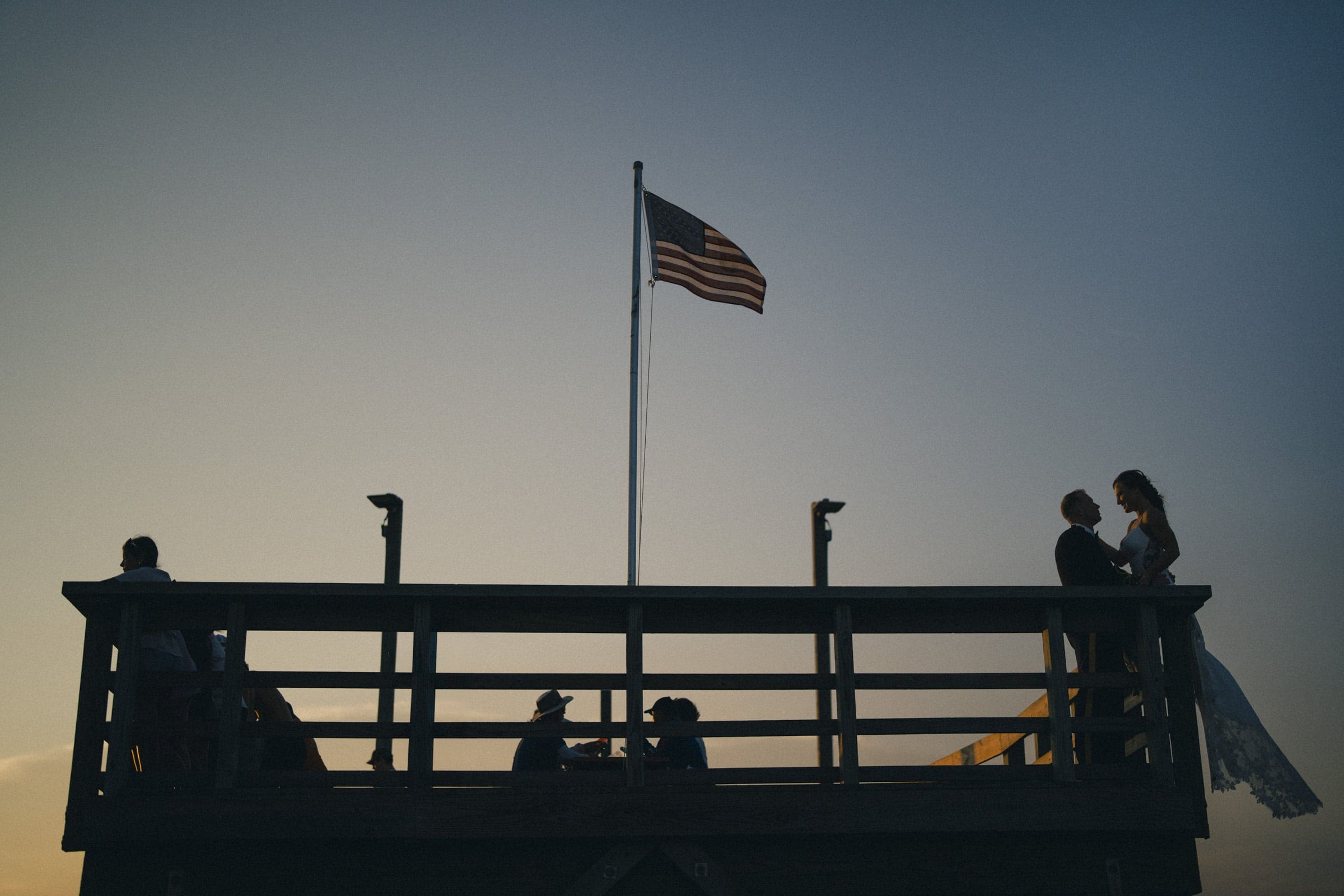 Silhouetted people stand and sit on a wooden platform with an American flag at the center, set against a dusky sky; a couple appears to be embracing or dancing on the right side.