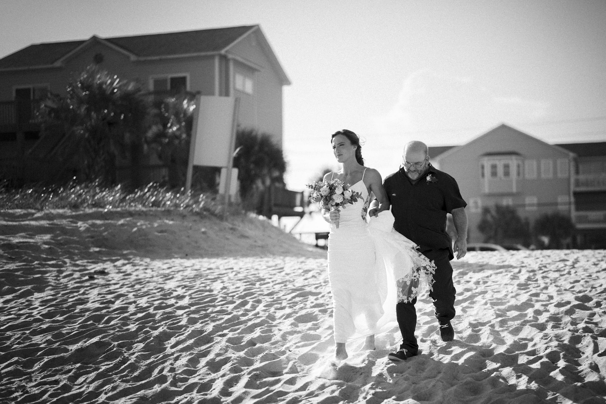 A bride in a white dress holding a bouquet walks on a sandy beach next to an older man in dark clothing. Houses and sunlight are visible in the background.