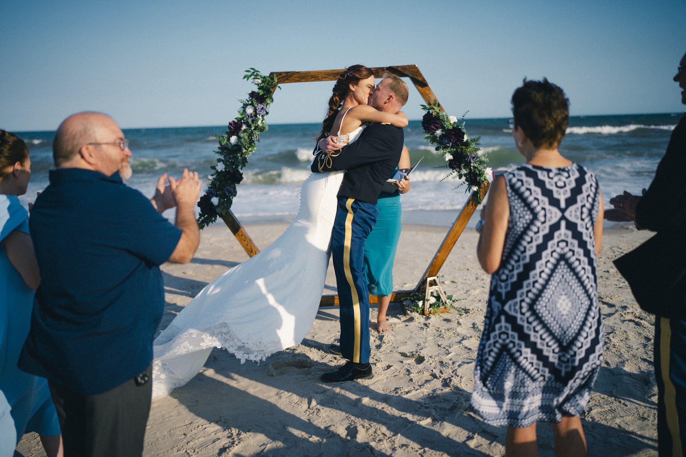 A bride and groom embrace and kiss under a wooden hexagon arch decorated with flowers on a sandy beach, while guests wearing blue outfits stand around them and applaud. Waves crash in the background under a clear sky.