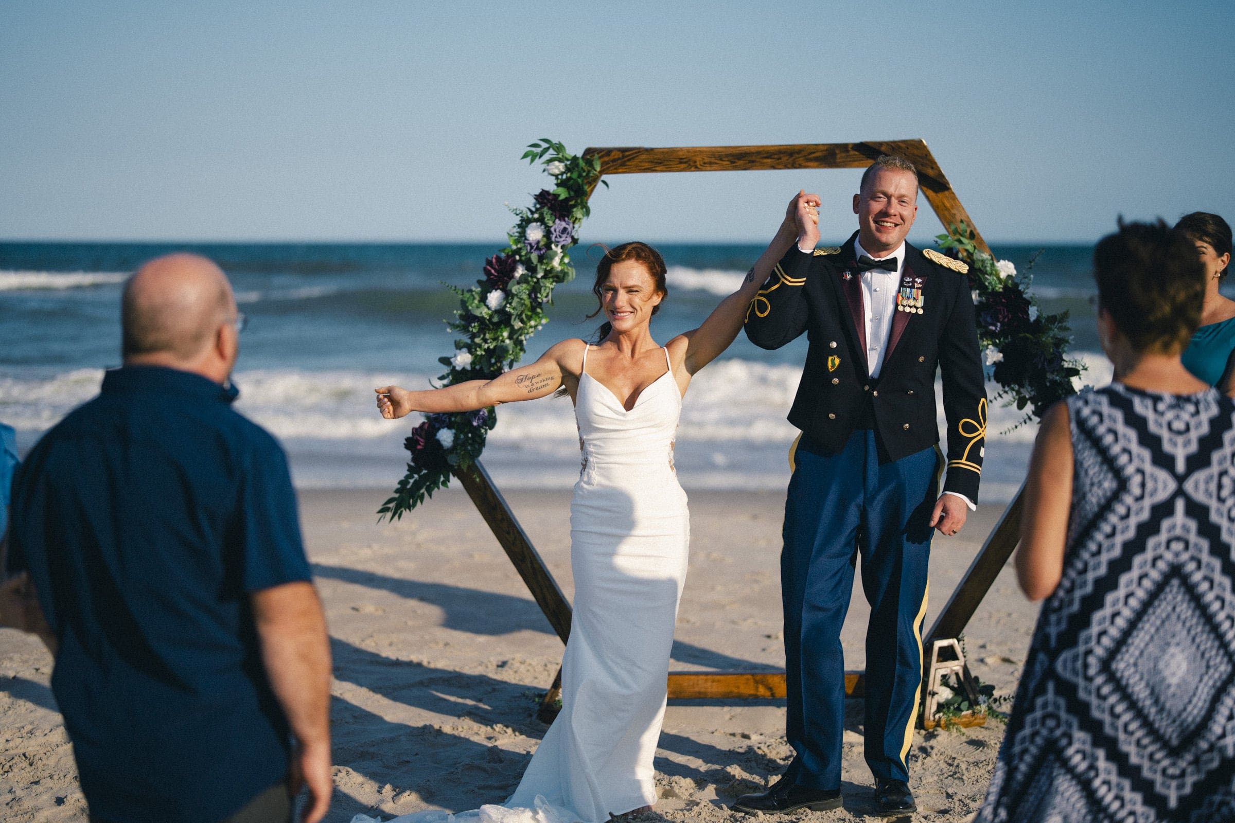A bride in a white dress and a groom in a military uniform hold hands and smile joyfully at a beach wedding, standing in front of a wooden hexagonal arch decorated with flowers, with guests watching.