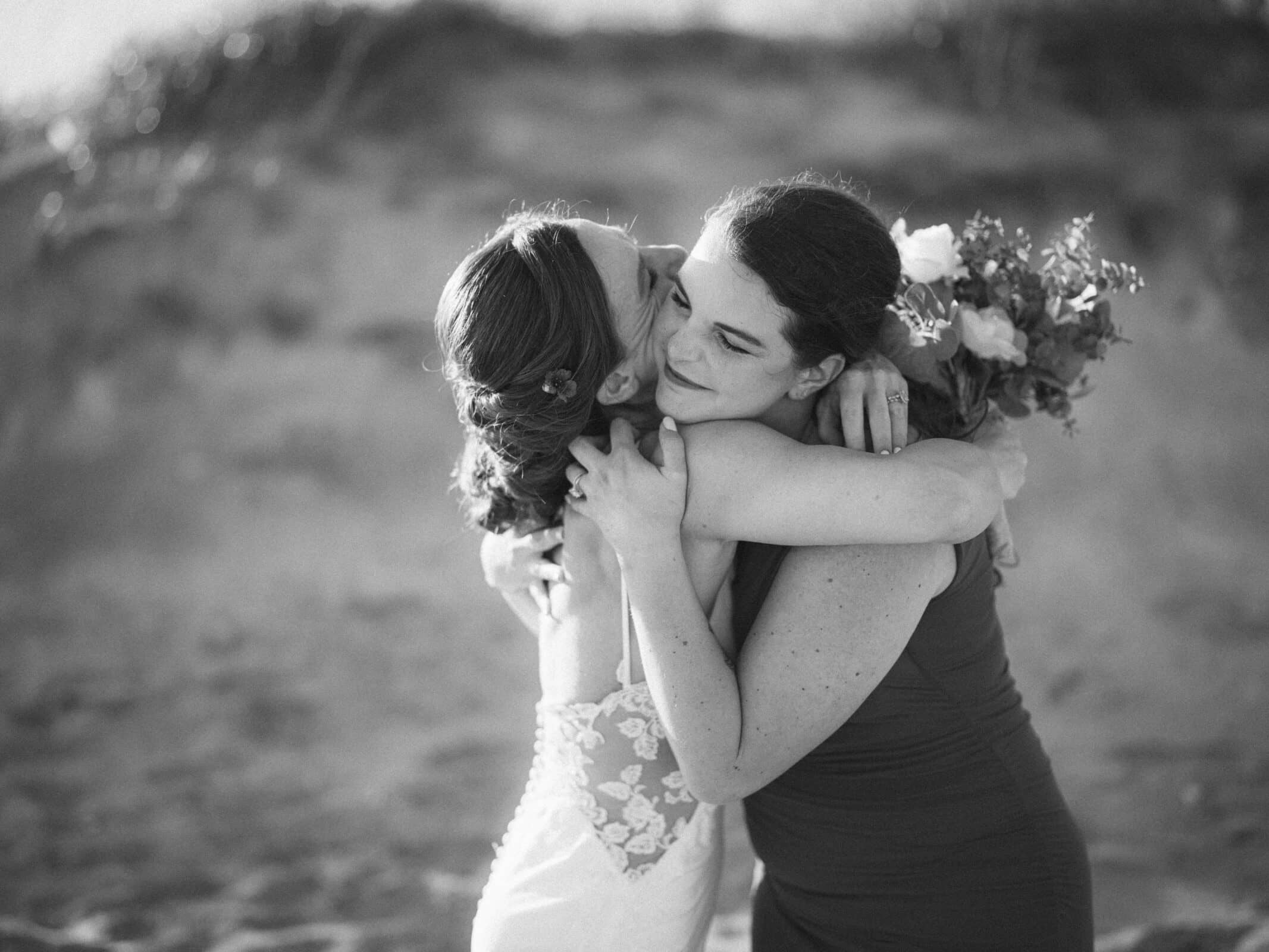 Two women hug closely on a beach, one wearing a wedding dress with lace details and the other holding a bouquet and wearing a dark dress. The moment appears joyful and emotional. The photo is in black and white.