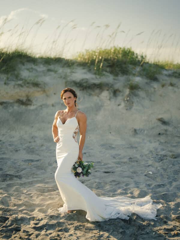 A woman in a white wedding dress stands on a sandy beach, holding a bouquet of flowers, with sunlight and tall grasses in the background.