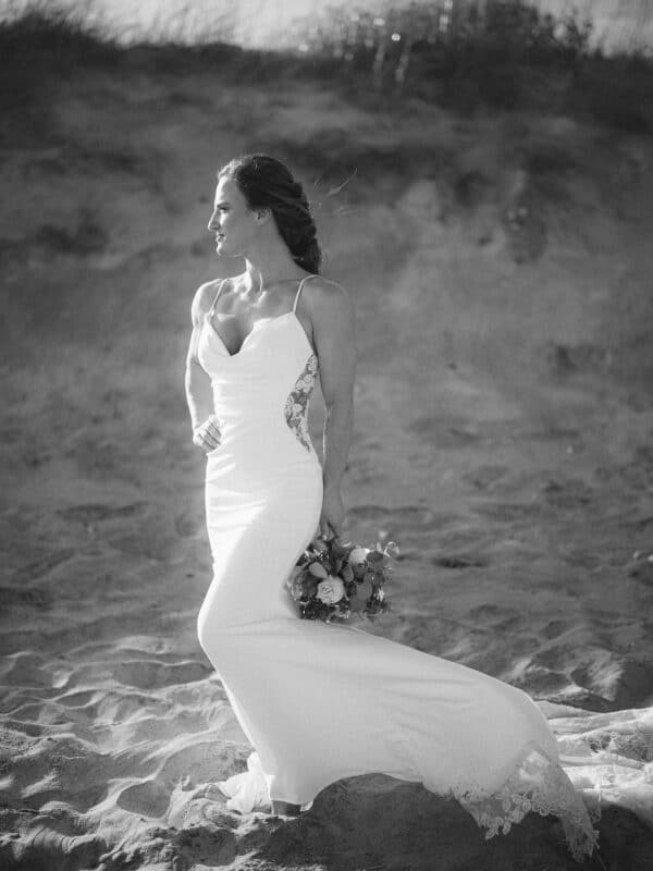 A woman in a white lace wedding dress stands on a sandy beach, holding a bouquet of flowers and looking to the side. The image is in black and white, with a blurred background of sand and grass.
