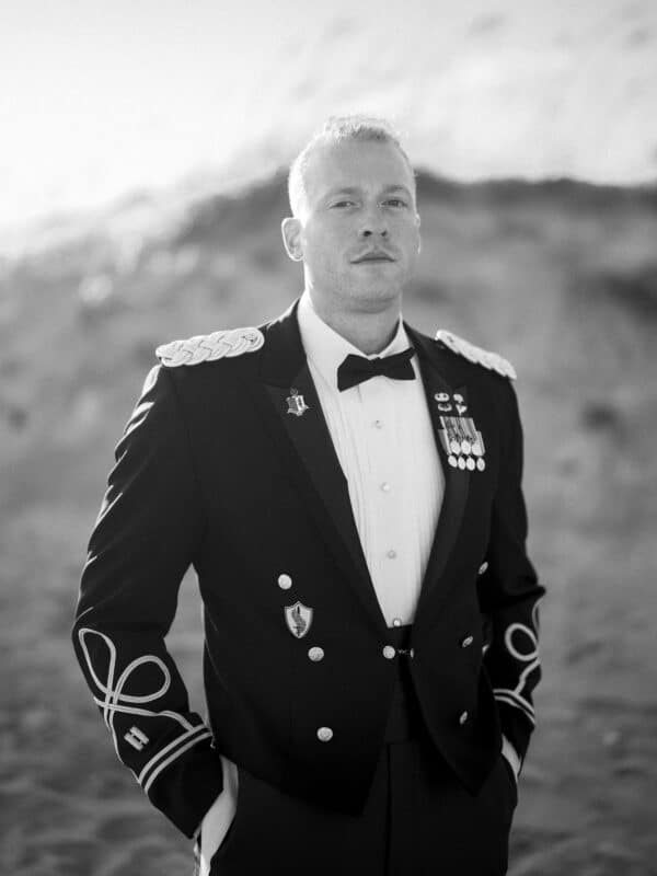 A man in formal military dress uniform stands outdoors with hands in pockets, looking confidently at the camera. He wears medals and insignia on his jacket. The background is blurred, suggesting an outdoor setting.