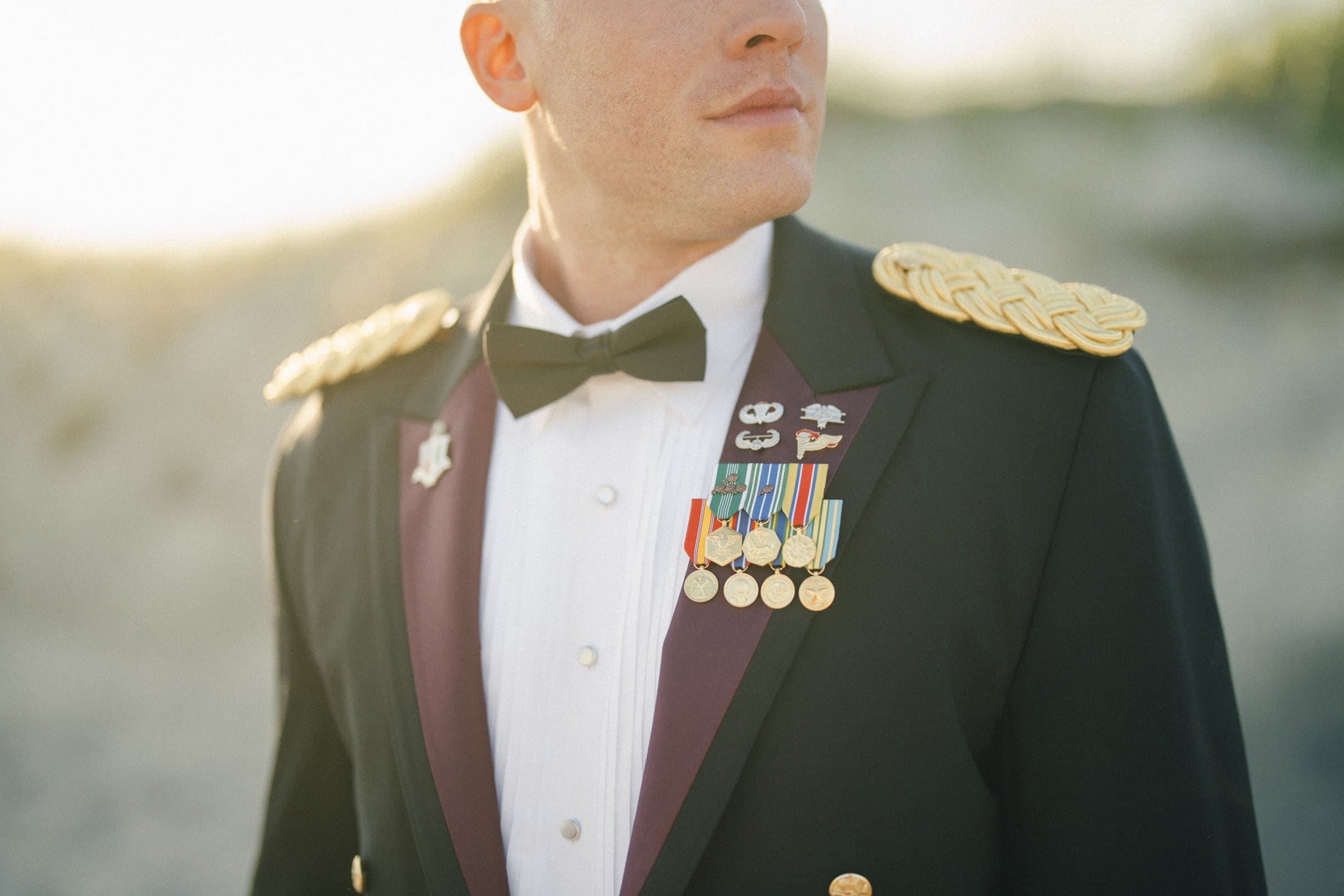 A man in a formal military dress uniform with gold shoulder decorations and several medals and ribbons stands outdoors, looking to the side. The background is blurred, suggesting a natural beach setting at sunset.