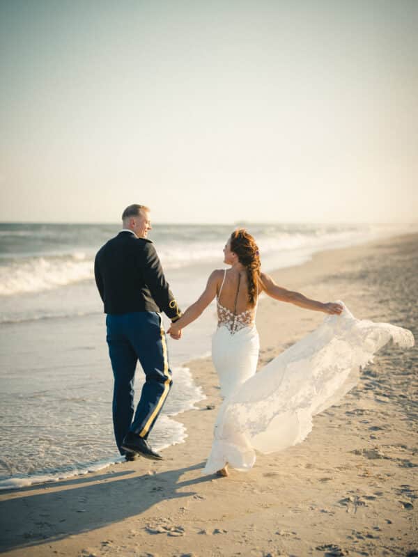 A bride in a white dress and a groom in a military uniform walk hand in hand along the beach at sunset, with waves in the background and her dress trailing in the sand.