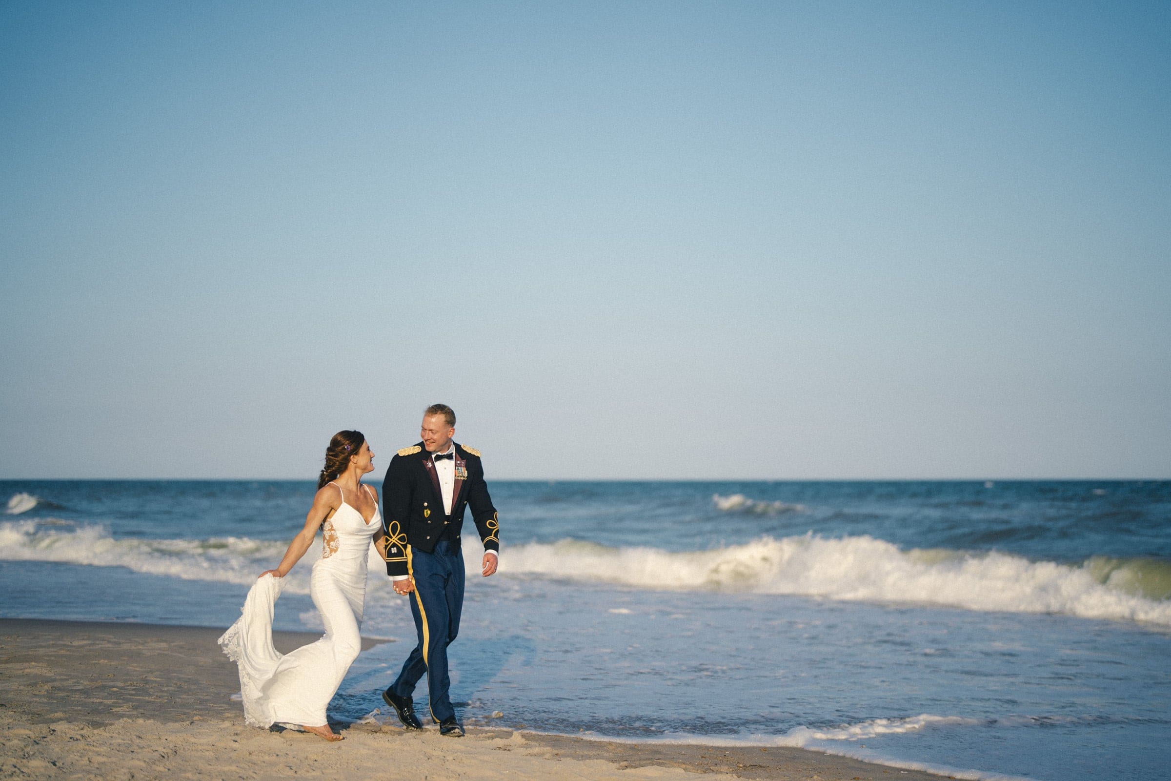 A bride in a white dress and a groom in a military uniform walk barefoot together on a sandy beach near the ocean, smiling at each other under a clear blue sky.